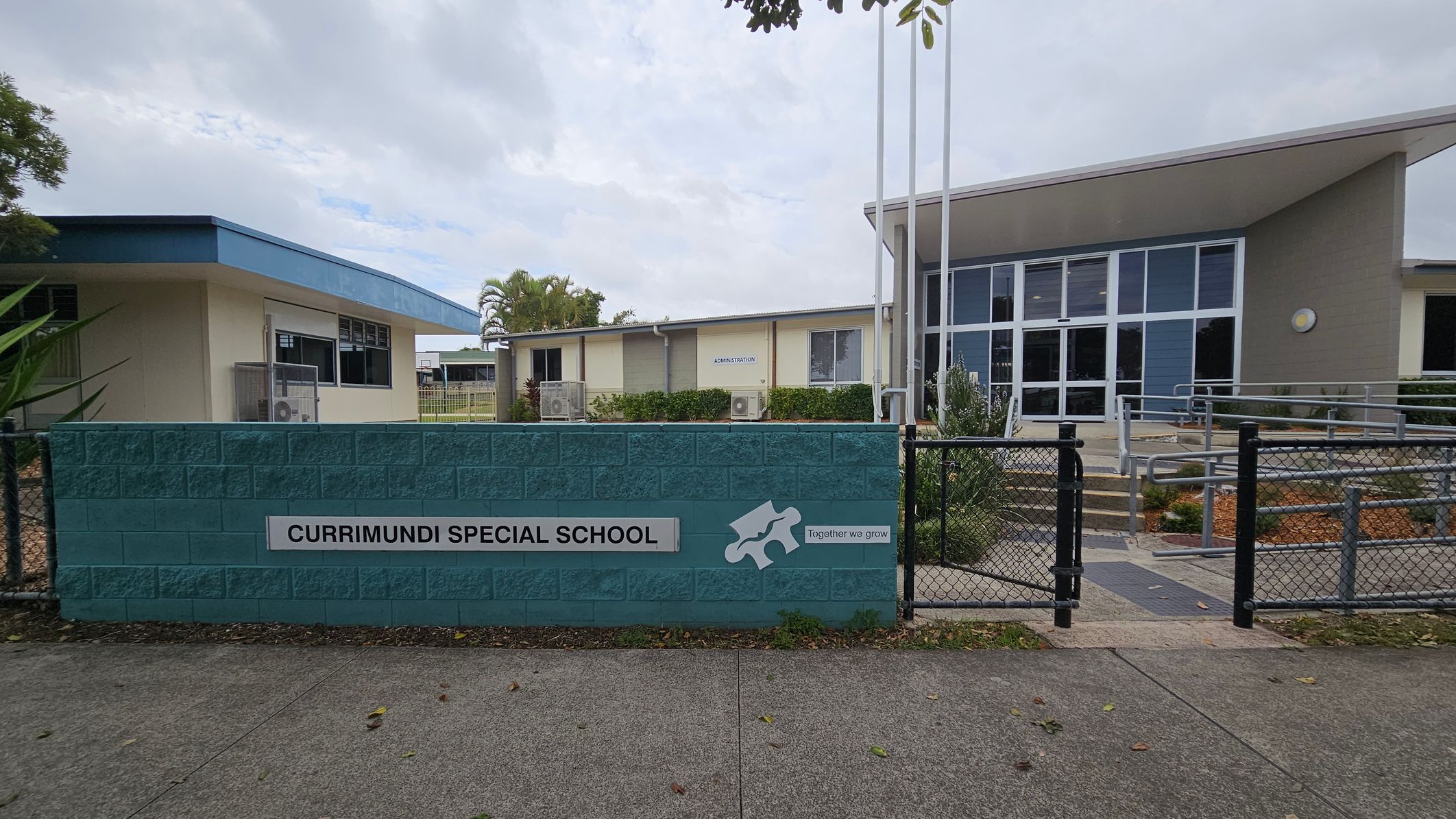 A school entrance with a blue brick wall in the foreground displaying the sign "Currimundi Special School" and the phrase "Together we grow" alongside a puzzle piece graphic. Behind the wall, there are two buildings. The left building has a light green exterior with a blue roof, while the right building features large glass windows and a sloped roof. A pathway with railings leads to the entrance. The ground is paved, and there are some plants and trees visible. A school entrance with a blue brick wall in the foreground displaying the sign "Currimundi Special School" and the phrase "Together we grow" alongside a puzzle piece graphic. Behind the wall, there are two buildings. The left building has a light green exterior with a blue roof, while the right building features large glass windows and a sloped roof. A pathway with railings leads to the entrance. The ground is paved, and there are some plants and trees visible.