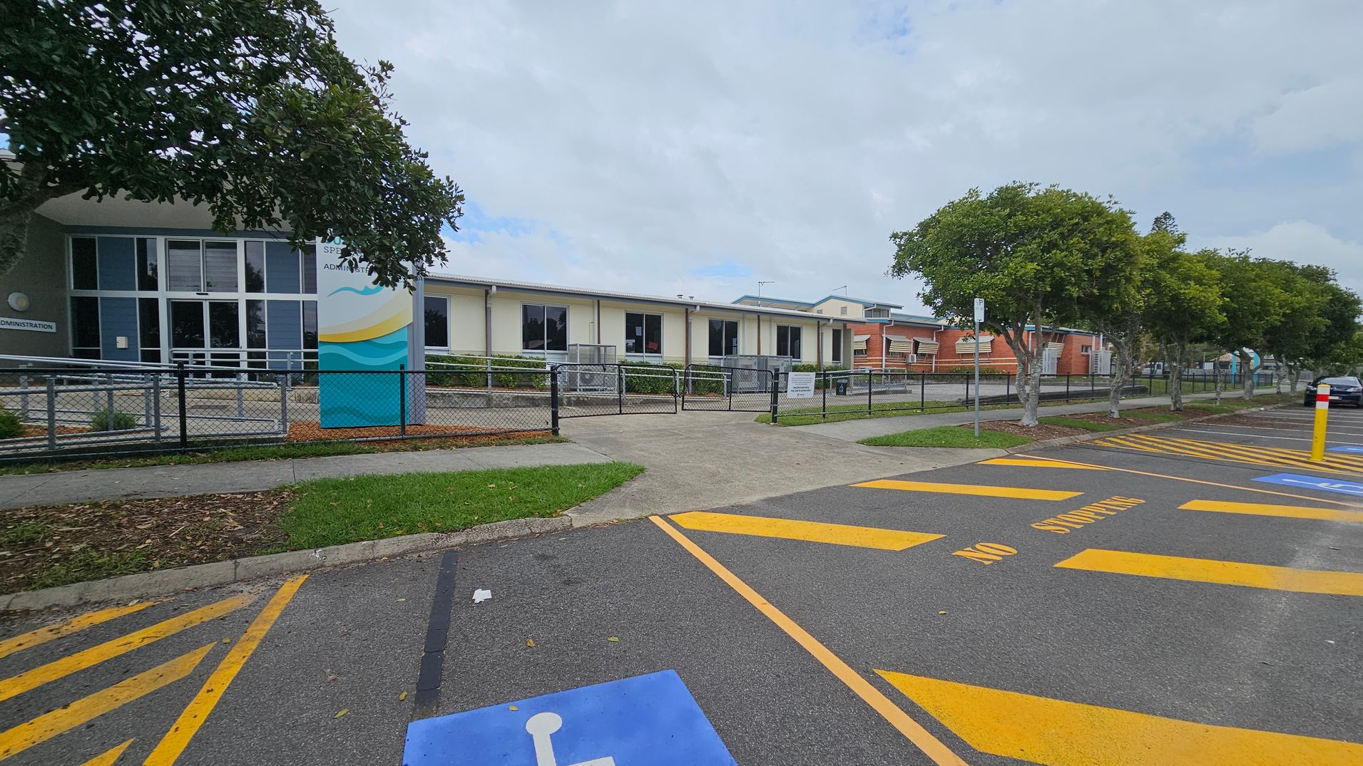 A school building with a modern design is in the background, featuring large windows and a sign near the entrance. A fence runs along the front, with a pathway leading to the entrance. Trees line the right side, providing some greenery. In the foreground, a car park is visible with marked spaces, including a blue disabled parking spot and yellow "No Stopping" lines. The ground is asphalt, and the area is well-maintained. A school building with a modern design is in the background, featuring large windows and a sign near the entrance. A fence runs along the front, with a pathway leading to the entrance. Trees line the right side, providing some greenery. In the foreground, a car park is visible with marked spaces, including a blue disabled parking spot and yellow "No Stopping" lines. The ground is asphalt, and the area is well-maintained.