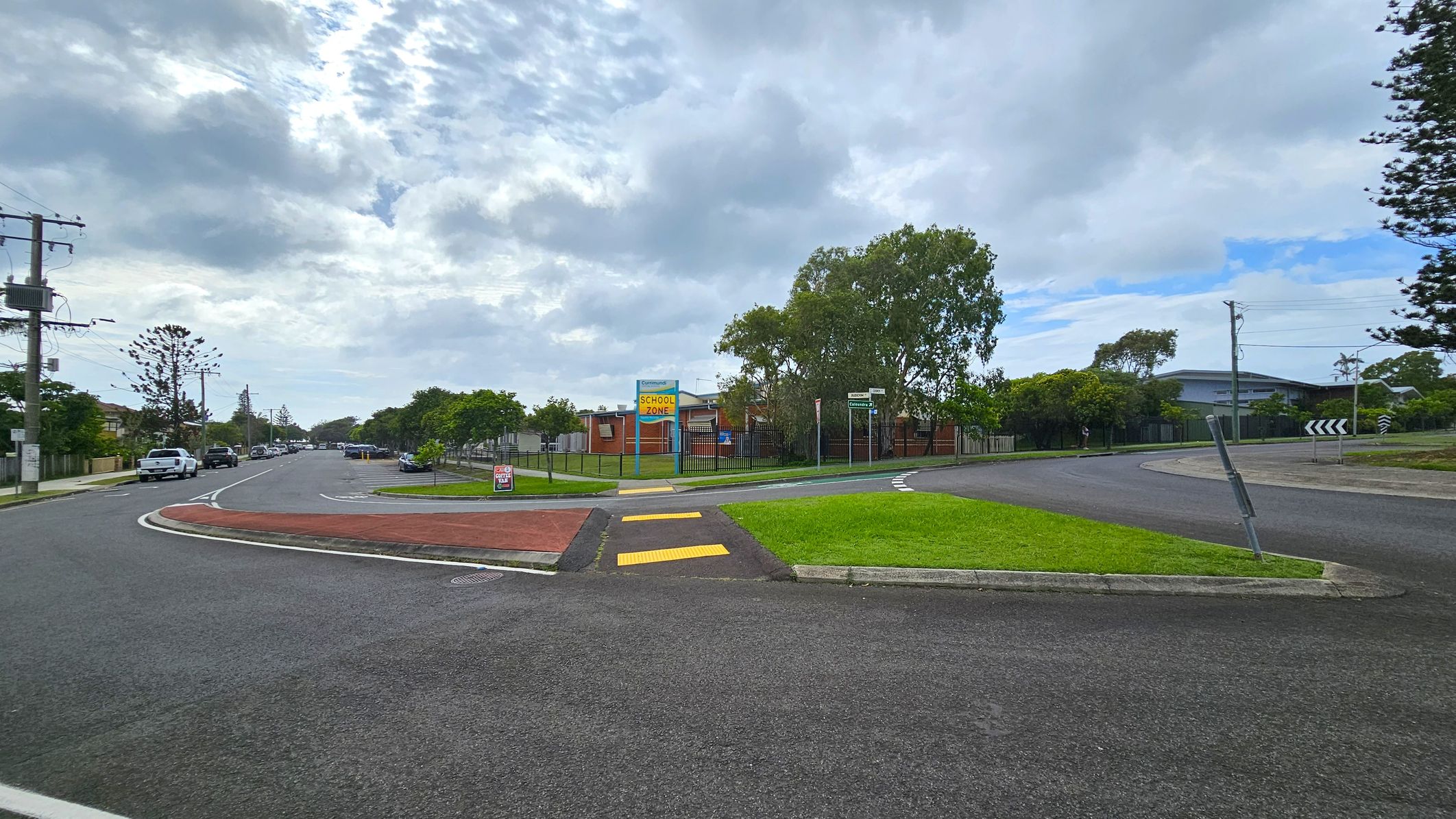 A street scene with a roundabout in the foreground. On the left, a road lined with parked cars and utility poles extends into the distance. In the centre, a grassy area with a "School Zone" sign and a pedestrian crossing with yellow markings. To the right, a curved road with a roundabout sign and trees in the background. A building is partially visible behind the trees. The sky is overcast with patches of blue. A street scene with a roundabout in the foreground. On the left, a road lined with parked cars and utility poles extends into the distance. In the centre, a grassy area with a "School Zone" sign and a pedestrian crossing with yellow markings. To the right, a curved road with a roundabout sign and trees in the background. A building is partially visible behind the trees. The sky is overcast with patches of blue.