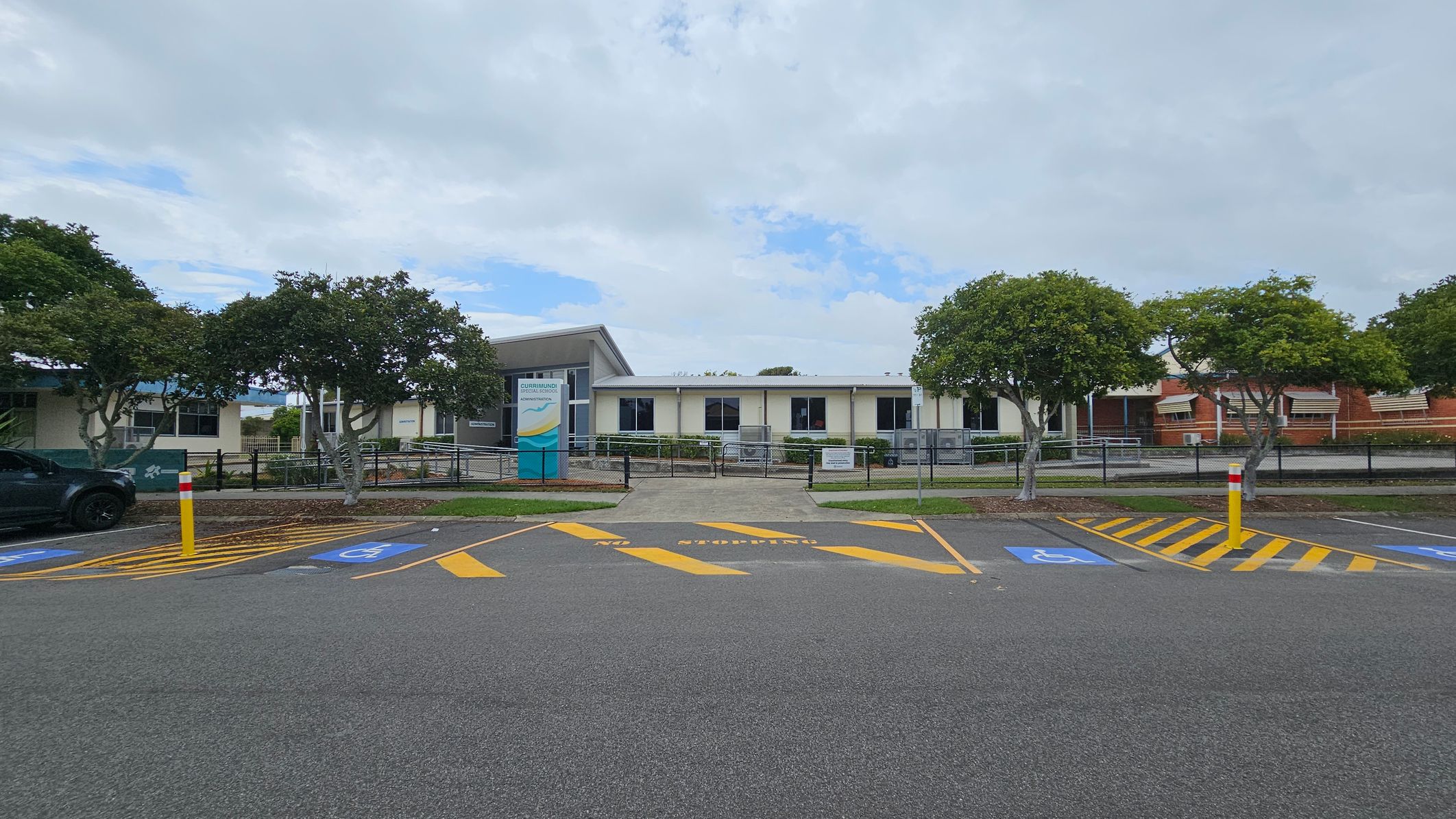 A single-storey building with a white facade and several windows is in the centre. In front, a sign with text is visible. The foreground features a car park with marked spaces, including disabled parking spots and yellow-striped areas. Trees with green foliage are on both sides, and a black fence runs along the front of the building. A grey asphalt surface covers the parking area. A single-storey building with a white facade and several windows is in the centre. In front, a sign with text is visible. The foreground features a car park with marked spaces, including disabled parking spots and yellow-striped areas. Trees with green foliage are on both sides, and a black fence runs along the front of the building. A grey asphalt surface covers the parking area.