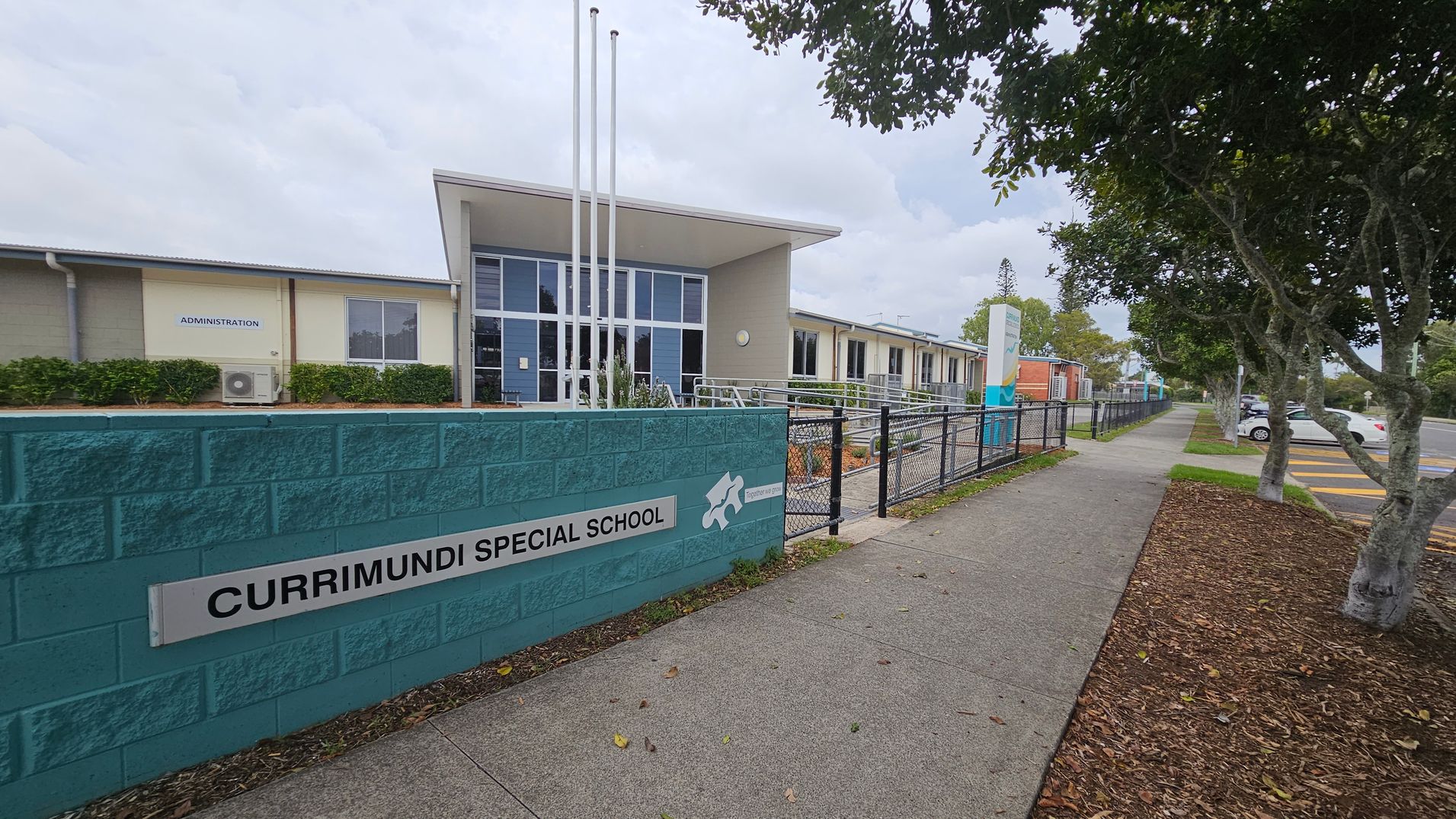 A school building with a modern design is on the left, featuring large windows and a sign reading "Administration." In front, a teal brick wall displays the name "Currimundi Special School." Three flagpoles stand nearby. On the right, a paved footpath runs alongside a fence, bordered by trees and a mulched area. Cars are parked in a lot further to the right. A school building with a modern design is on the left, featuring large windows and a sign reading "Administration." In front, a teal brick wall displays the name "Currimundi Special School." Three flagpoles stand nearby. On the right, a paved footpath runs alongside a fence, bordered by trees and a mulched area. Cars are parked in a lot further to the right.