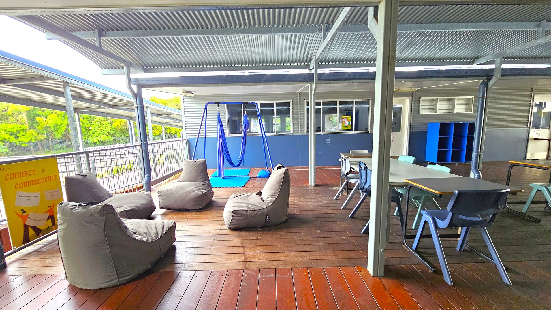 An outdoor covered area with a corrugated metal roof and wooden flooring. On the left, there are several grey bean bags and a yellow sign with text. In the centre, a blue swing hangs from a metal frame, with a blue mat underneath. On the right, there are tables and chairs, and a blue shelving unit against the wall. The background features a building with windows and a railing, with greenery visible beyond. An outdoor covered area with a corrugated metal roof and wooden flooring. On the left, there are several grey bean bags and a yellow sign with text. In the centre, a blue swing hangs from a metal frame, with a blue mat underneath. On the right, there are tables and chairs, and a blue shelving unit against the wall. The background features a building with windows and a railing, with greenery visible beyond.