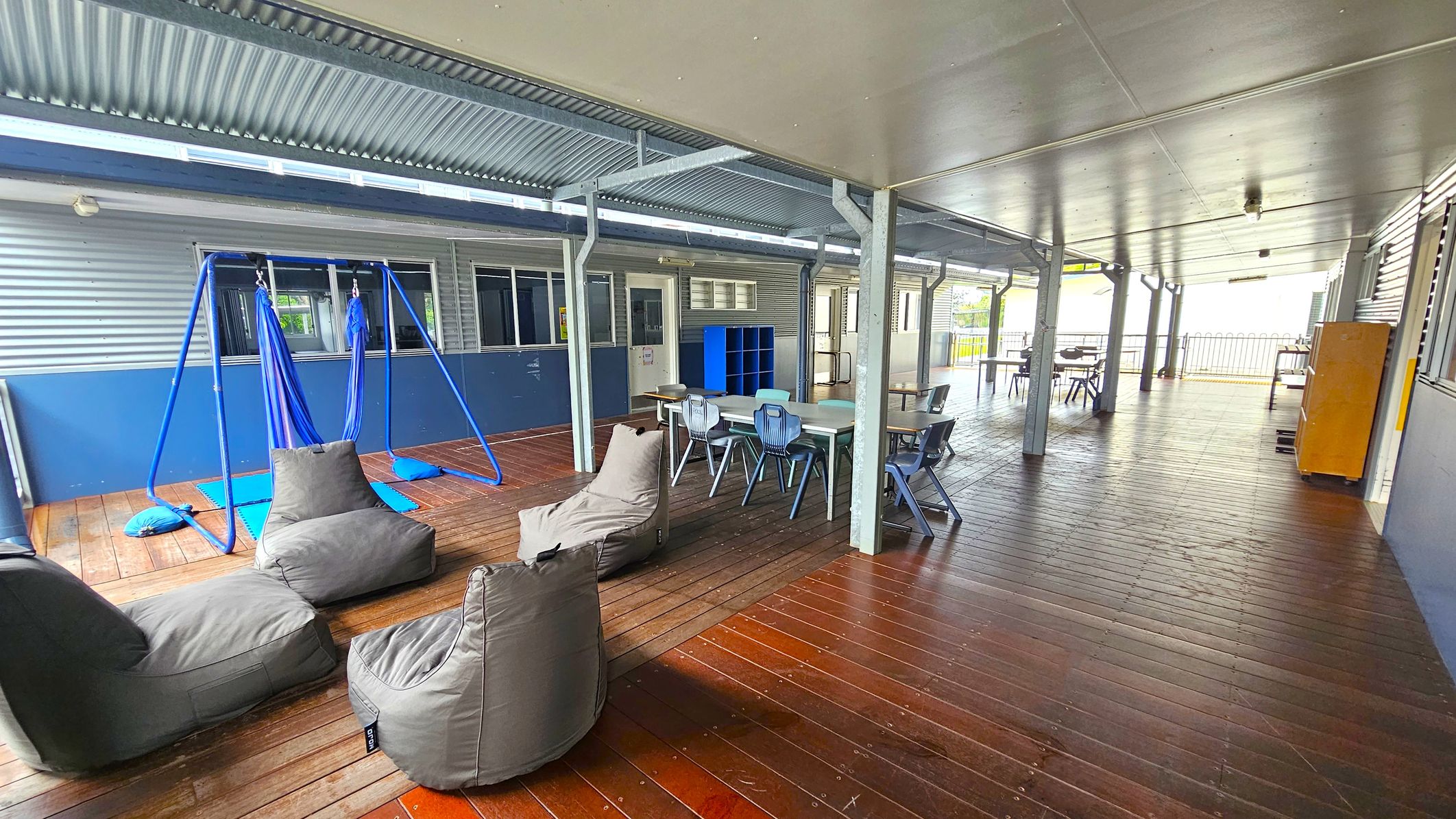 A covered outdoor area with a wooden floor and a metal ceiling. On the left, there are grey bean bags and a blue swing set with fabric seats. In the middle, a table with several blue chairs is positioned near a blue shelving unit. The right side features more tables and chairs, with a yellow cabinet against the wall. The background shows a corrugated metal wall with windows and doors, and a railing at the far end. A covered outdoor area with a wooden floor and a metal ceiling. On the left, there are grey bean bags and a blue swing set with fabric seats. In the middle, a table with several blue chairs is positioned near a blue shelving unit. The right side features more tables and chairs, with a yellow cabinet against the wall. The background shows a corrugated metal wall with windows and doors, and a railing at the far end.