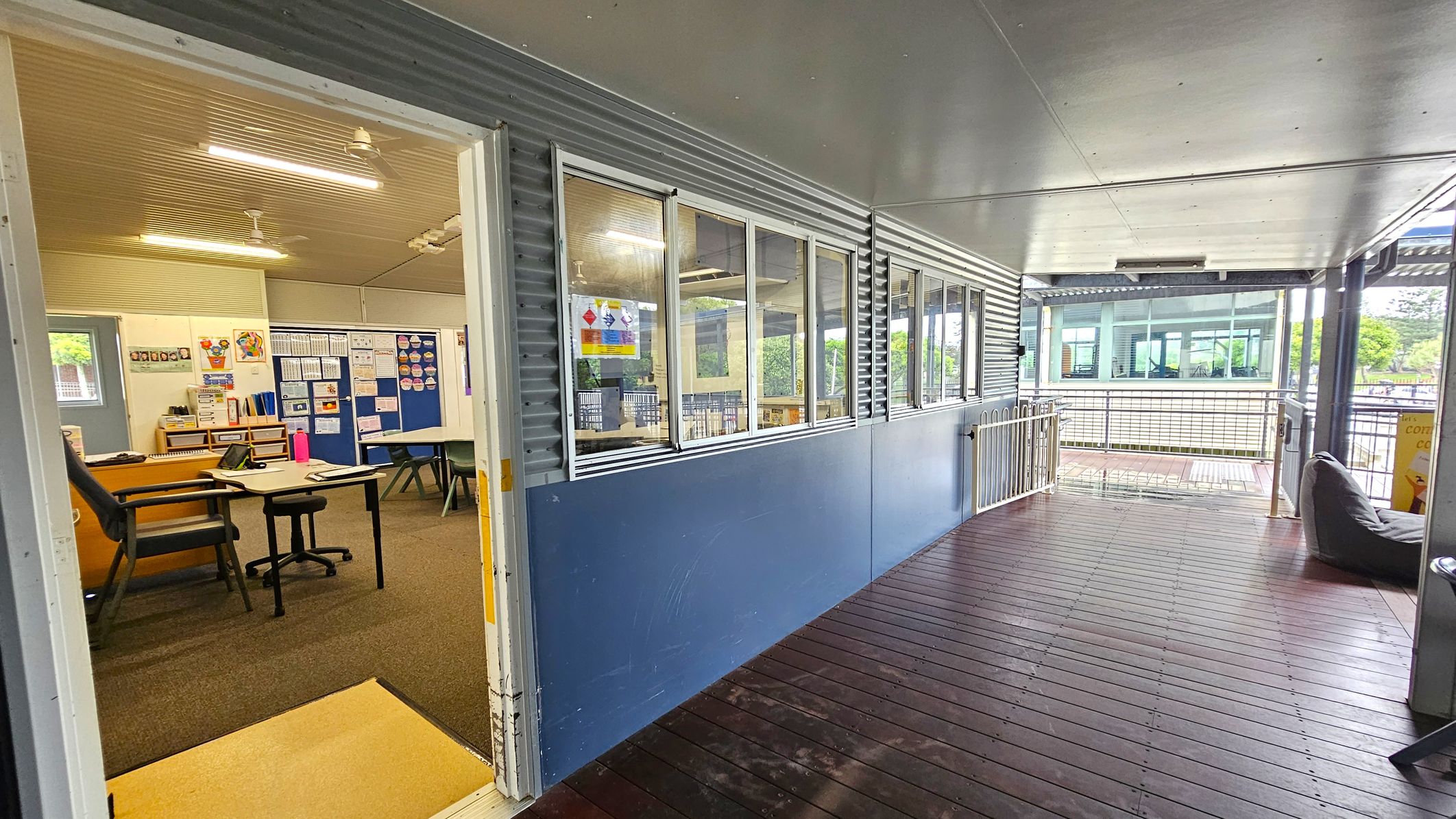 A corridor with a wooden floor and a metal ceiling. On the left, an open door leads to a classroom with a desk, chair, and educational posters on the walls. The classroom has a carpeted floor. The right side of the corridor features large windows with a blue lower wall. In the background, there is a railing and a view of another building with large windows and greenery visible outside. A corridor with a wooden floor and a metal ceiling. On the left, an open door leads to a classroom with a desk, chair, and educational posters on the walls. The classroom has a carpeted floor. The right side of the corridor features large windows with a blue lower wall. In the background, there is a railing and a view of another building with large windows and greenery visible outside.