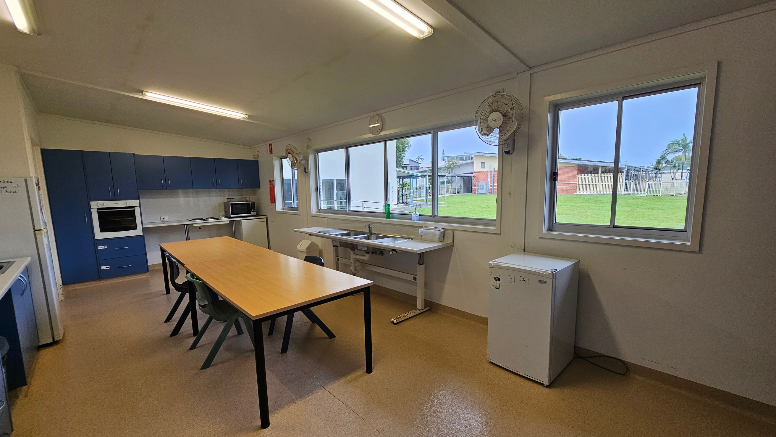 A kitchen with blue cabinets on the left, featuring an oven and a microwave. A long wooden table with black chairs is in the centre. Large windows on the right provide a view of a grassy area and buildings outside. Below the windows are two sinks and a small white fridge. The ceiling has fluorescent lights, and the floor is a light brown linoleum. Two wall-mounted fans are above the windows. A kitchen with blue cabinets on the left, featuring an oven and a microwave. A long wooden table with black chairs is in the centre. Large windows on the right provide a view of a grassy area and buildings outside. Below the windows are two sinks and a small white fridge. The ceiling has fluorescent lights, and the floor is a light brown linoleum. Two wall-mounted fans are above the windows.