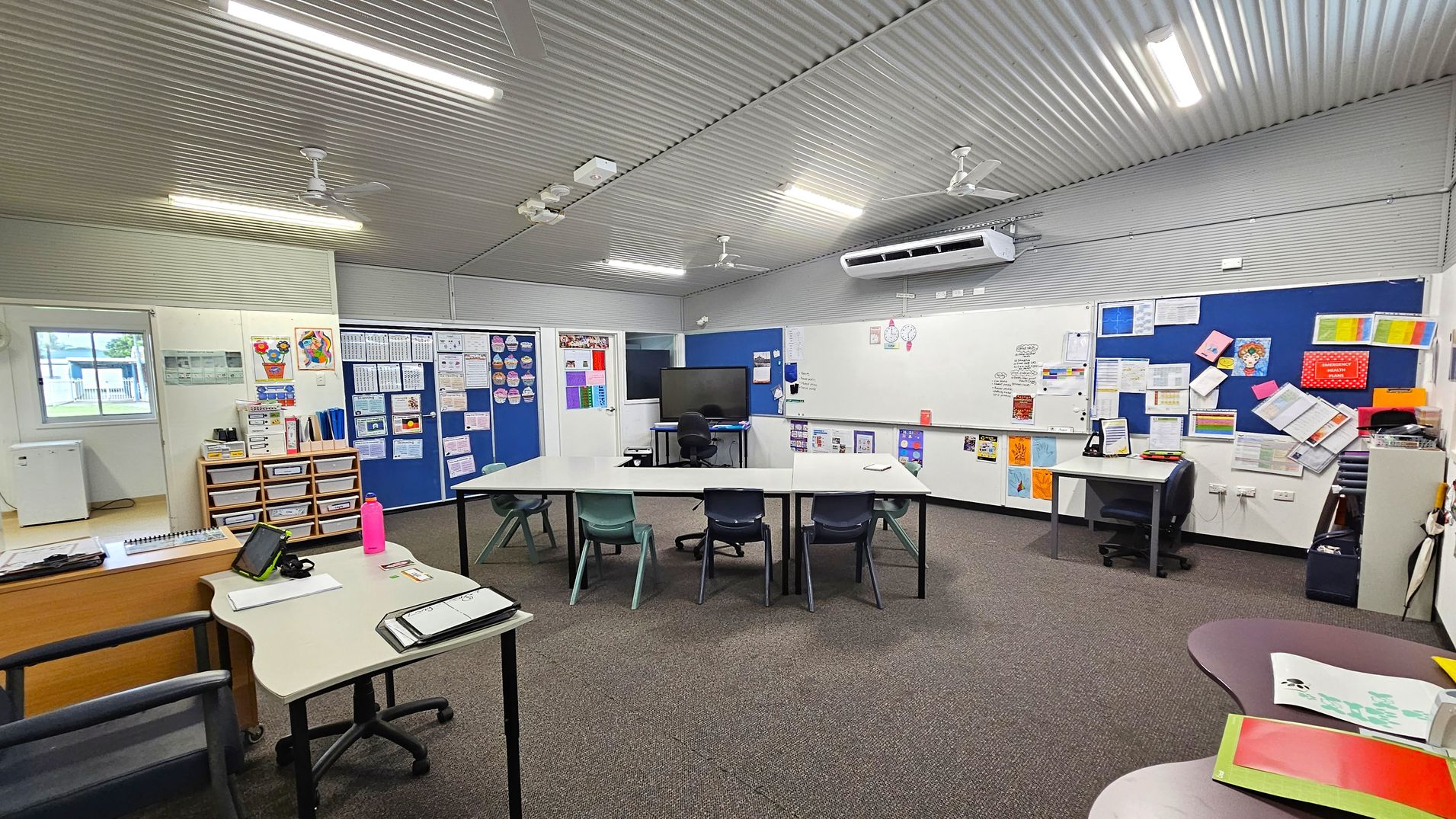 A classroom with a corrugated metal ceiling and fluorescent lights. The floor is carpeted. In the centre, there are tables arranged in a U-shape with various chairs. On the right, a large whiteboard and blue noticeboards are filled with papers and colourful materials. A desk with a chair and a monitor is also present. On the left, a window, storage drawers, and a small desk with a tablet and a pink water bottle. The walls are decorated with educational posters and charts. A classroom with a corrugated metal ceiling and fluorescent lights. The floor is carpeted. In the centre, there are tables arranged in a U-shape with various chairs. On the right, a large whiteboard and blue noticeboards are filled with papers and colourful materials. A desk with a chair and a monitor is also present. On the left, a window, storage drawers, and a small desk with a tablet and a pink water bottle. The walls are decorated with educational posters and charts.