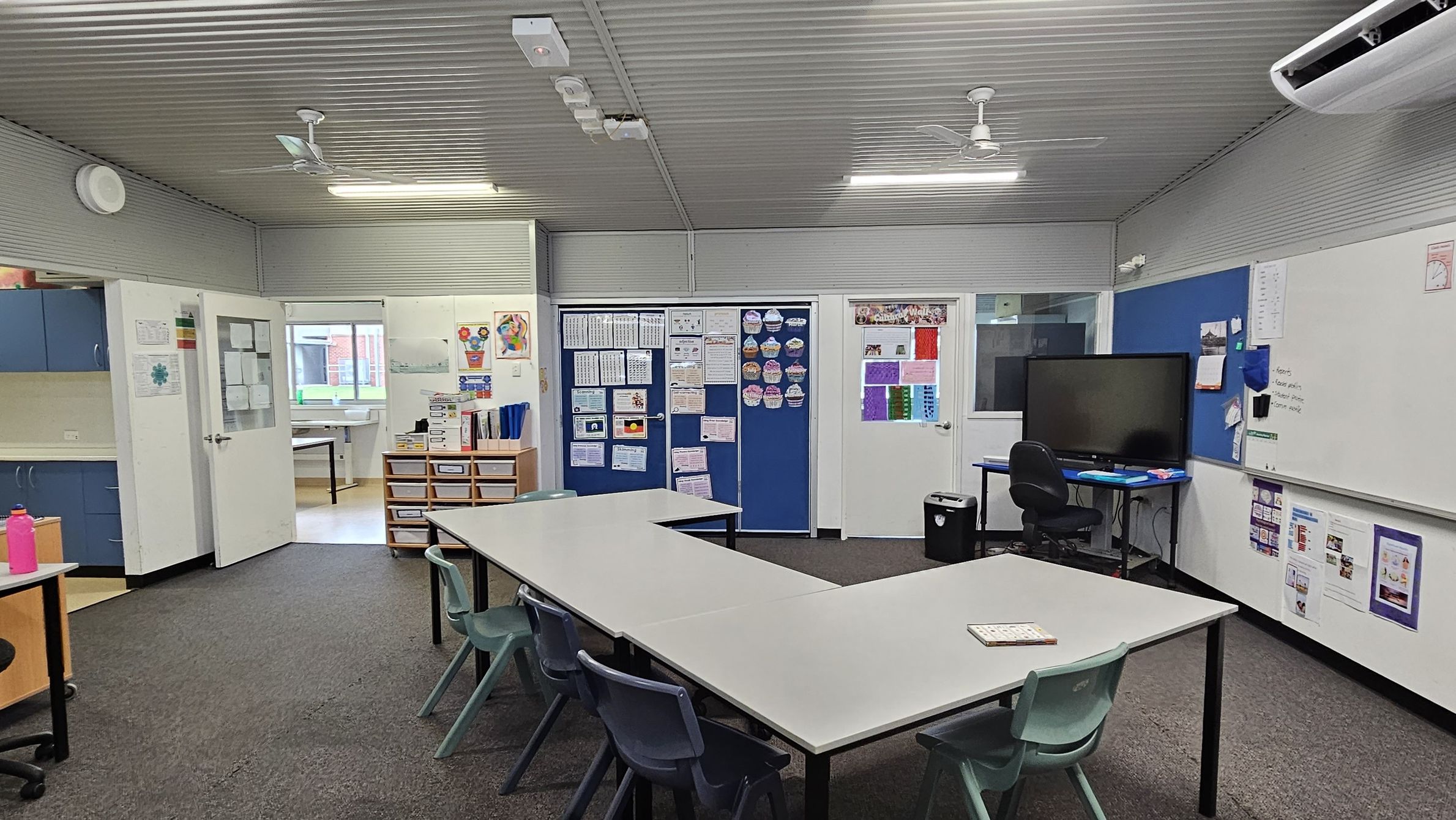 A classroom with a U-shaped arrangement of white tables and green chairs in the centre. On the right, there's a large whiteboard and a blue noticeboard with a flat-screen monitor on a desk. The back wall features blue and white doors with educational posters. On the left, blue cabinets and a pink water bottle are visible. The floor is carpeted, and the ceiling is corrugated with fluorescent lights and a ceiling fan. An air conditioning unit is mounted on the right wall. A classroom with a U-shaped arrangement of white tables and green chairs in the centre. On the right, there's a large whiteboard and a blue noticeboard with a flat-screen monitor on a desk. The back wall features blue and white doors with educational posters. On the left, blue cabinets and a pink water bottle are visible. The floor is carpeted, and the ceiling is corrugated with fluorescent lights and a ceiling fan. An air conditioning unit is mounted on the right wall.