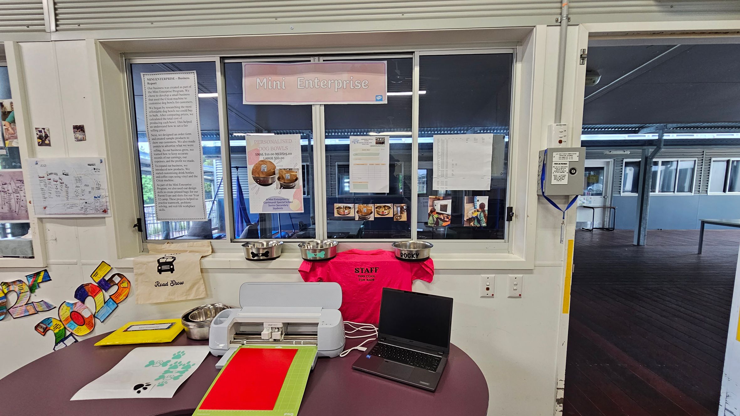 A room with a window labelled "Mini Enterprise" displaying various notices and posters. Below the window, there are several metal dog bowls, a bright pink staff T-shirt, and a laptop on a round table. The table also holds a cutting machine, colourful paper, and a sheet with paw prints. On the left wall, there are colourful cut-out numbers and a small poster. The floor is wooden, and the ceiling has visible beams. The background shows an open area with more windows and a wooden floor. A room with a window labelled "Mini Enterprise" displaying various notices and posters. Below the window, there are several metal dog bowls, a bright pink staff T-shirt, and a laptop on a round table. The table also holds a cutting machine, colourful paper, and a sheet with paw prints. On the left wall, there are colourful cut-out numbers and a small poster. The floor is wooden, and the ceiling has visible beams. The background shows an open area with more windows and a wooden floor.
