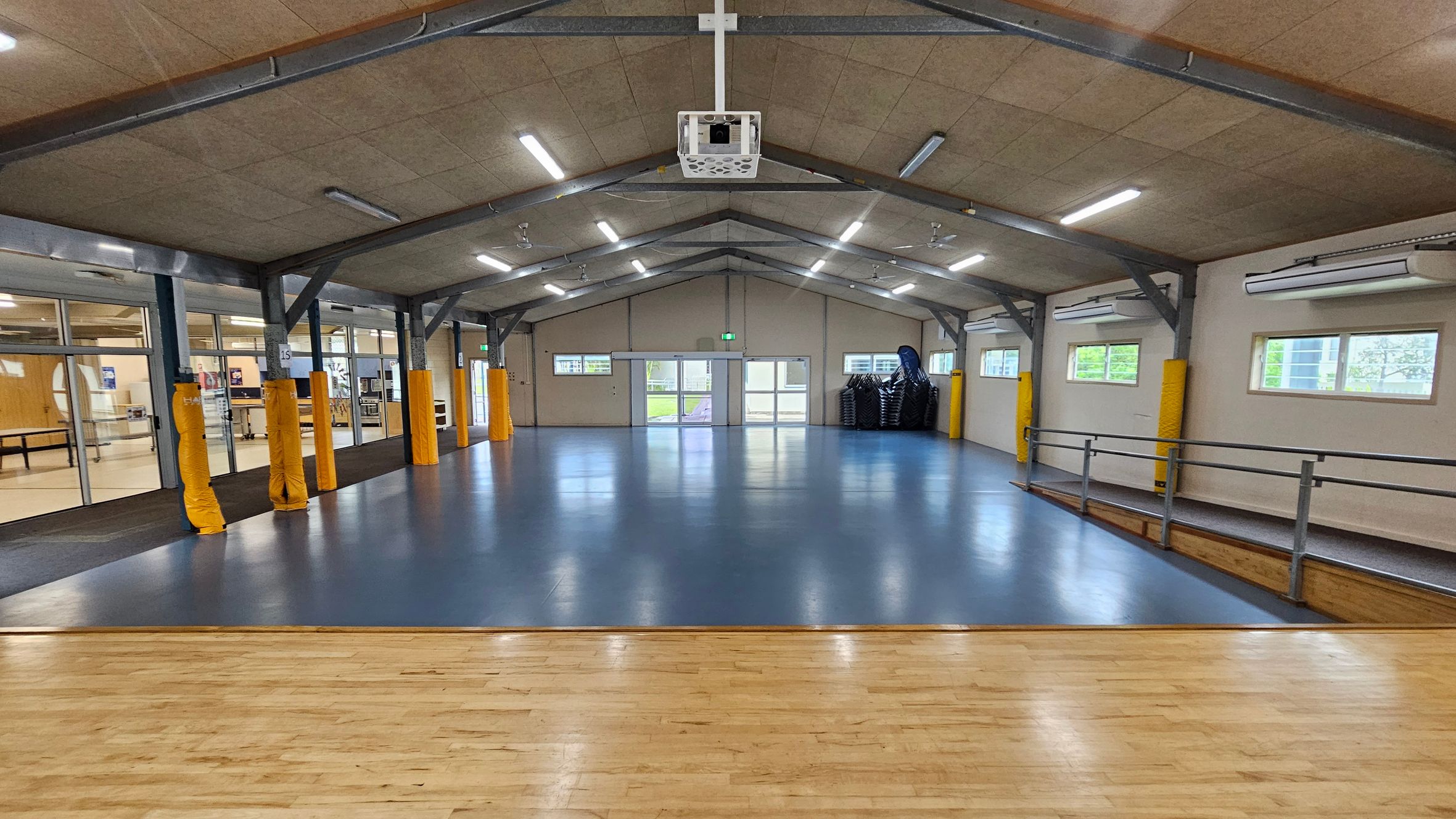 A spacious hall with a blue floor and a wooden stage in the foreground. The ceiling is high with exposed beams and fluorescent lights. On the left, there are large glass windows and doors leading to another room. On the right, windows allow natural light in, and a stack of chairs is visible. The back wall has double doors and small windows. Yellow padding covers some structural beams. The hall has a clean, open appearance. A spacious hall with a blue floor and a wooden stage in the foreground. The ceiling is high with exposed beams and fluorescent lights. On the left, there are large glass windows and doors leading to another room. On the right, windows allow natural light in, and a stack of chairs is visible. The back wall has double doors and small windows. Yellow padding covers some structural beams. The hall has a clean, open appearance.
