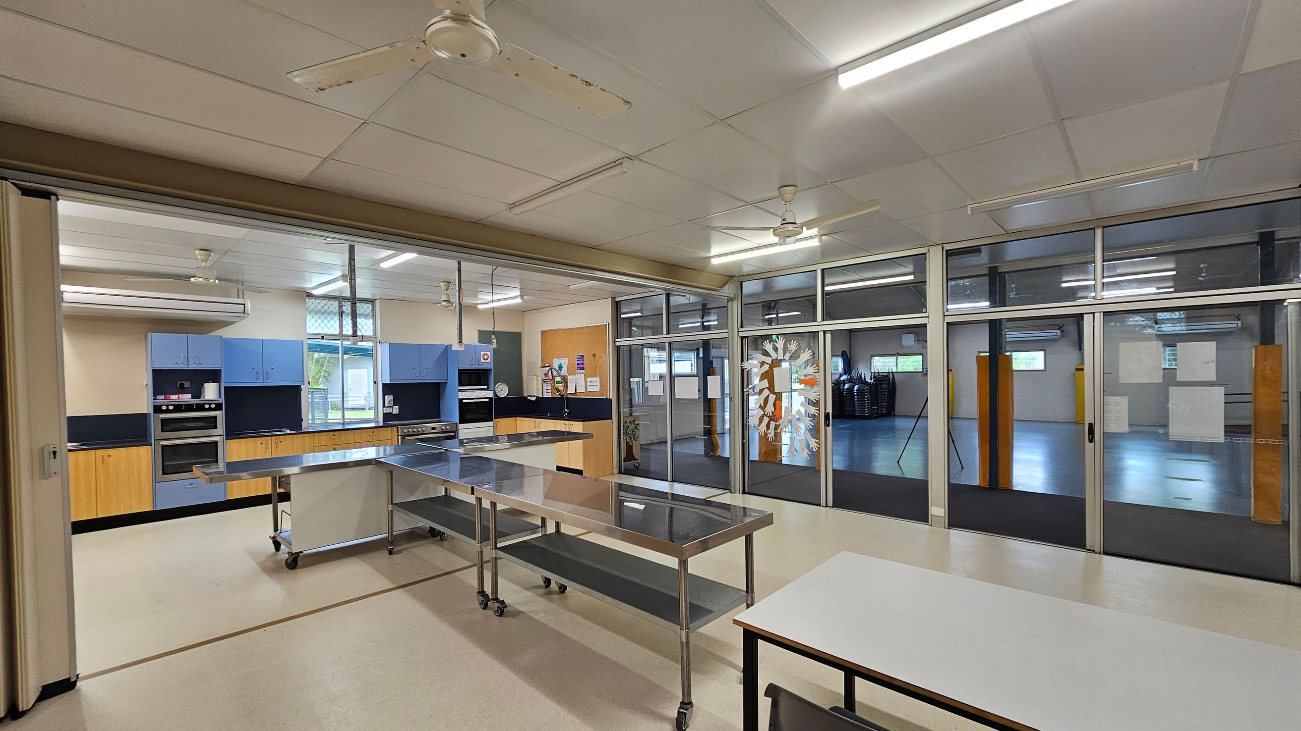A spacious kitchen area with light-coloured flooring and a ceiling with fluorescent lights and ceiling fans. On the left, there are blue cabinets and a double oven. In the centre, a stainless steel table on wheels is positioned. The right side features large glass doors with paper decorations, leading to an adjacent room with stacked chairs and more windows. A white table is in the foreground. A spacious kitchen area with light-coloured flooring and a ceiling with fluorescent lights and ceiling fans. On the left, there are blue cabinets and a double oven. In the centre, a stainless steel table on wheels is positioned. The right side features large glass doors with paper decorations, leading to an adjacent room with stacked chairs and more windows. A white table is in the foreground.