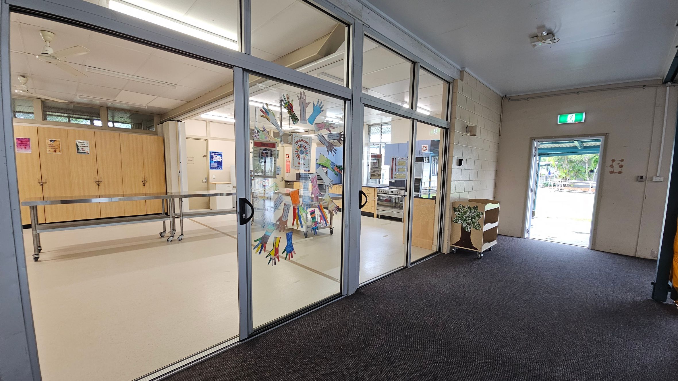 A room with glass sliding doors decorated with colourful handprints. Inside, there are wooden cabinets and a metal table on wheels. The ceiling has fluorescent lights and a ceiling fan. The floor is carpeted in the foreground and tiled inside the room. To the right, an open door leads outside, with a small shelf nearby. Posters are visible on the walls and cabinets. A room with glass sliding doors decorated with colourful handprints. Inside, there are wooden cabinets and a metal table on wheels. The ceiling has fluorescent lights and a ceiling fan. The floor is carpeted in the foreground and tiled inside the room. To the right, an open door leads outside, with a small shelf nearby. Posters are visible on the walls and cabinets.