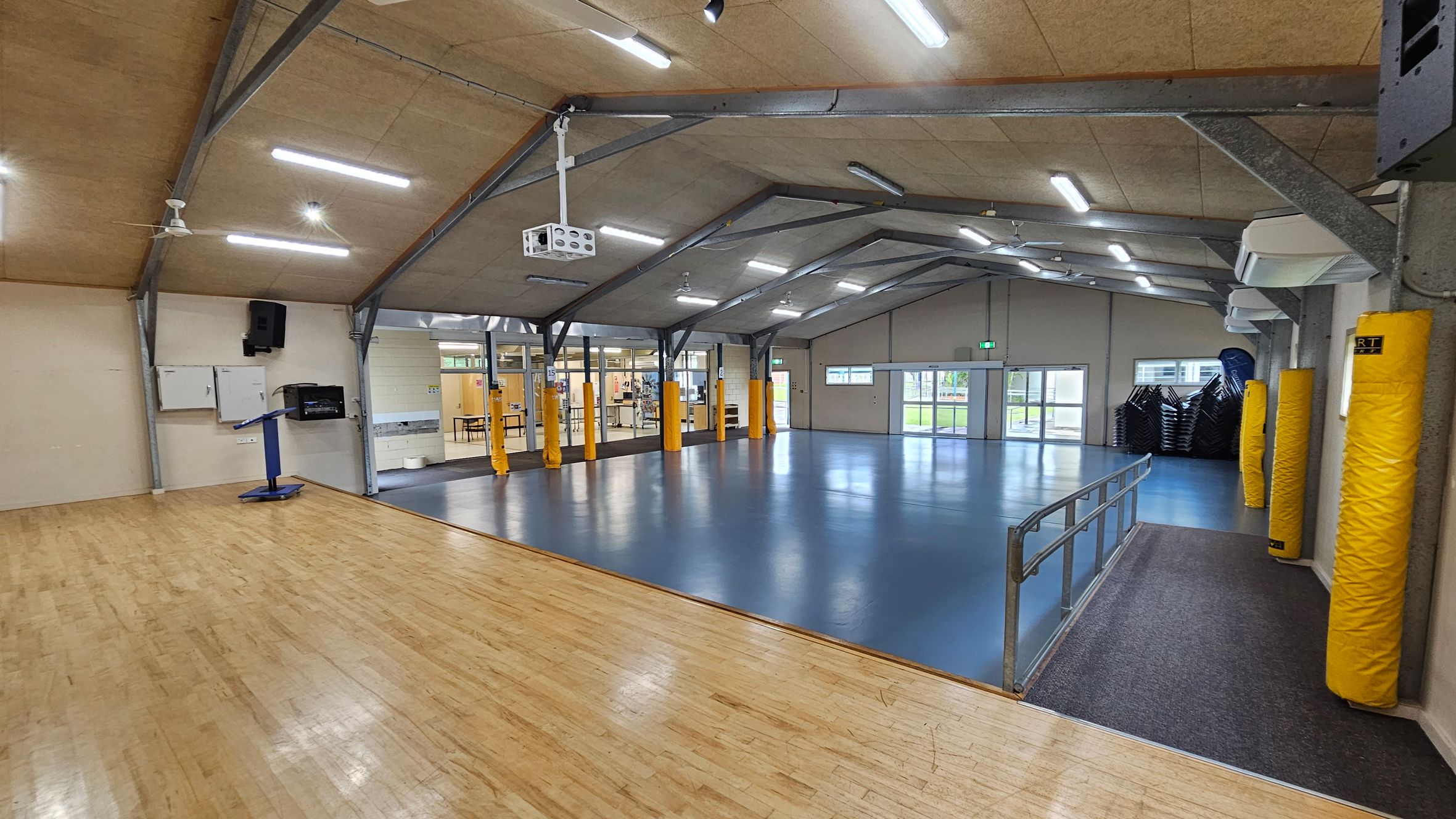 A spacious indoor hall with a wooden floor on the left and a blue floor in the centre and right. The ceiling is high with exposed beams and fluorescent lights. Yellow padded columns are evenly spaced along the walls. A projector is mounted on the ceiling. On the right, there are stacked chairs near a set of double doors. A metal railing separates the wooden and blue floor areas. The background features glass doors and windows leading to another room. A spacious indoor hall with a wooden floor on the left and a blue floor in the centre and right. The ceiling is high with exposed beams and fluorescent lights. Yellow padded columns are evenly spaced along the walls. A projector is mounted on the ceiling. On the right, there are stacked chairs near a set of double doors. A metal railing separates the wooden and blue floor areas. The background features glass doors and windows leading to another room.