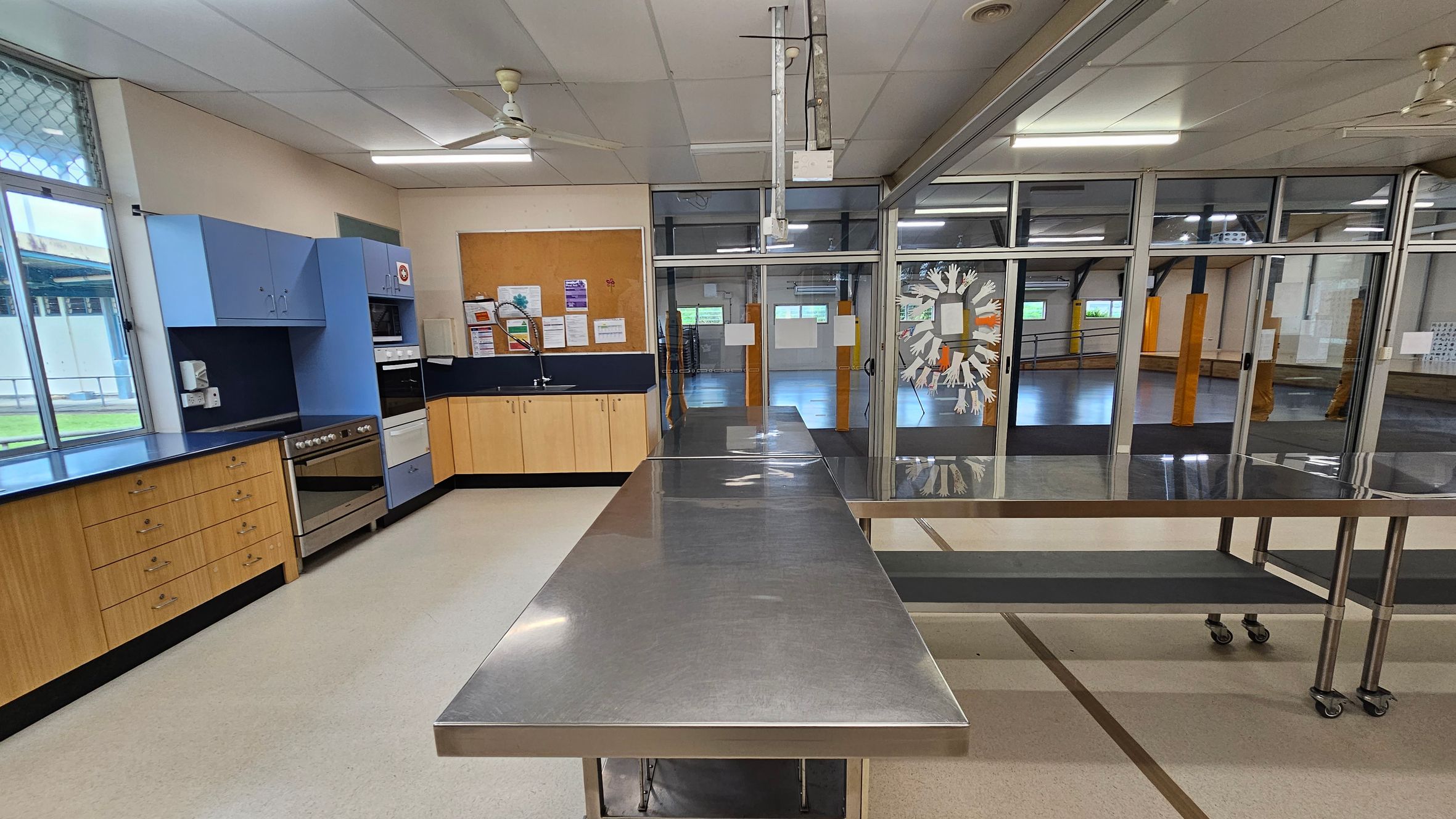 A kitchen with light-coloured flooring and a white ceiling with fluorescent lights and ceiling fans. On the left, there are wooden cabinets with a blue countertop, a stainless steel oven, and a microwave. A window with a mesh screen is above the counter. In the centre, a long stainless steel table on wheels. The right side features glass doors with a decorative design, leading to a large open room with visible support beams and windows. A kitchen with light-coloured flooring and a white ceiling with fluorescent lights and ceiling fans. On the left, there are wooden cabinets with a blue countertop, a stainless steel oven, and a microwave. A window with a mesh screen is above the counter. In the centre, a long stainless steel table on wheels. The right side features glass doors with a decorative design, leading to a large open room with visible support beams and windows.