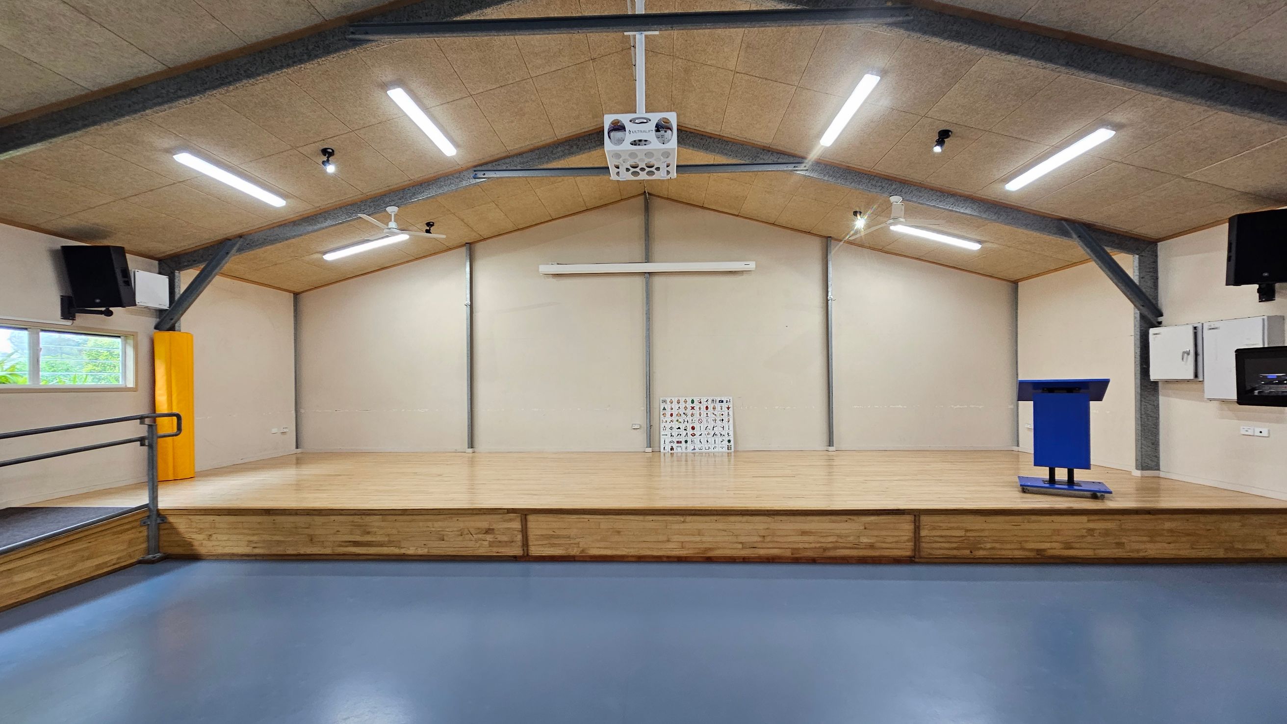 A spacious room with a wooden stage in the centre, featuring a blue podium on the right. The floor is blue, and the ceiling has exposed beams with several fluorescent lights. On the left, there's a window with a yellow padded column nearby. The back wall has a small chart with various symbols or letters. Two black speakers are mounted on either side of the room. A spacious room with a wooden stage in the centre, featuring a blue podium on the right. The floor is blue, and the ceiling has exposed beams with several fluorescent lights. On the left, there's a window with a yellow padded column nearby. The back wall has a small chart with various symbols or letters. Two black speakers are mounted on either side of the room.