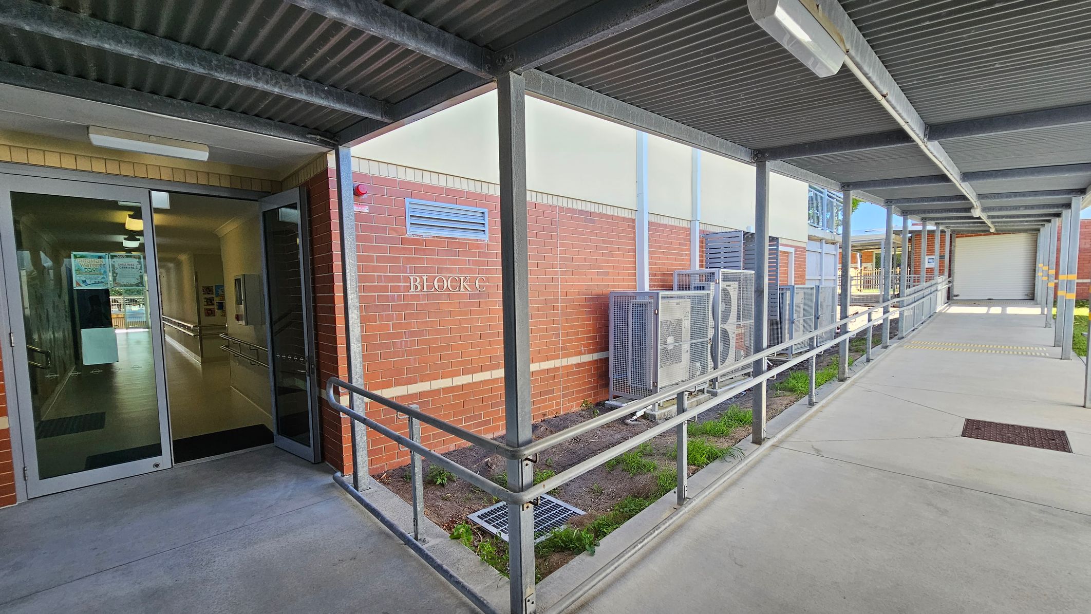 A covered walkway with a metal roof and railings leads to a building entrance on the left. The entrance has glass doors, and inside, a hallway is visible. The building is made of red brick with a sign reading "BLOCK C." Air conditioning units are mounted on the exterior wall. The floor is concrete, and there is a small patch of grass and soil beside the walkway. The ceiling has fluorescent lights. A covered walkway with a metal roof and railings leads to a building entrance on the left. The entrance has glass doors, and inside, a hallway is visible. The building is made of red brick with a sign reading "BLOCK C." Air conditioning units are mounted on the exterior wall. The floor is concrete, and there is a small patch of grass and soil beside the walkway. The ceiling has fluorescent lights.