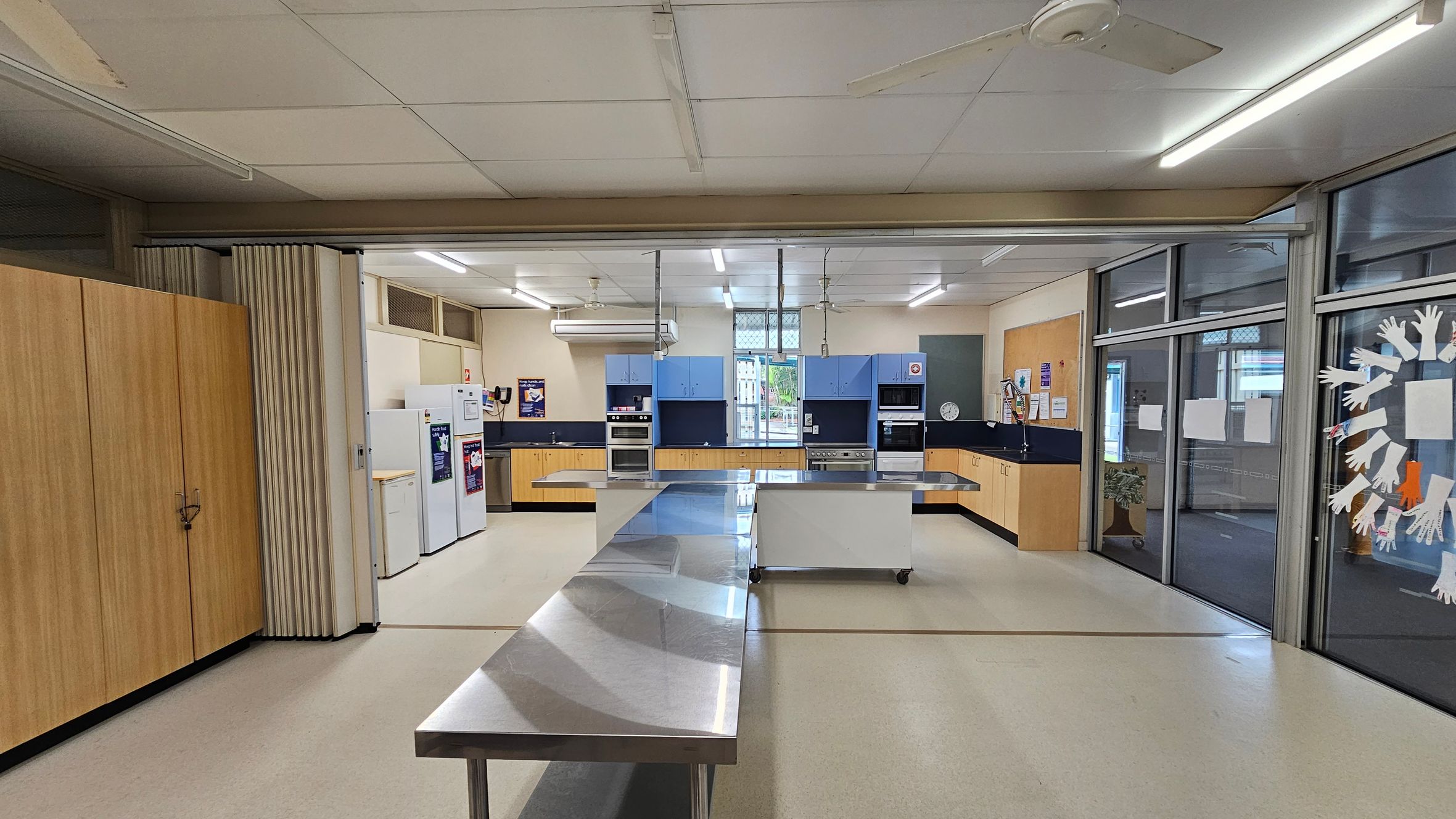 A spacious kitchen with a central stainless steel workbench. On the left, there are wooden cabinets and a row of white appliances, including fridges. The back wall features blue and wooden cabinetry with built-in ovens and a sink. On the right, large glass windows display paper cut-outs of hands. The ceiling has fluorescent lights and a fan, and the floor is a light-coloured linoleum. A spacious kitchen with a central stainless steel workbench. On the left, there are wooden cabinets and a row of white appliances, including fridges. The back wall features blue and wooden cabinetry with built-in ovens and a sink. On the right, large glass windows display paper cut-outs of hands. The ceiling has fluorescent lights and a fan, and the floor is a light-coloured linoleum.