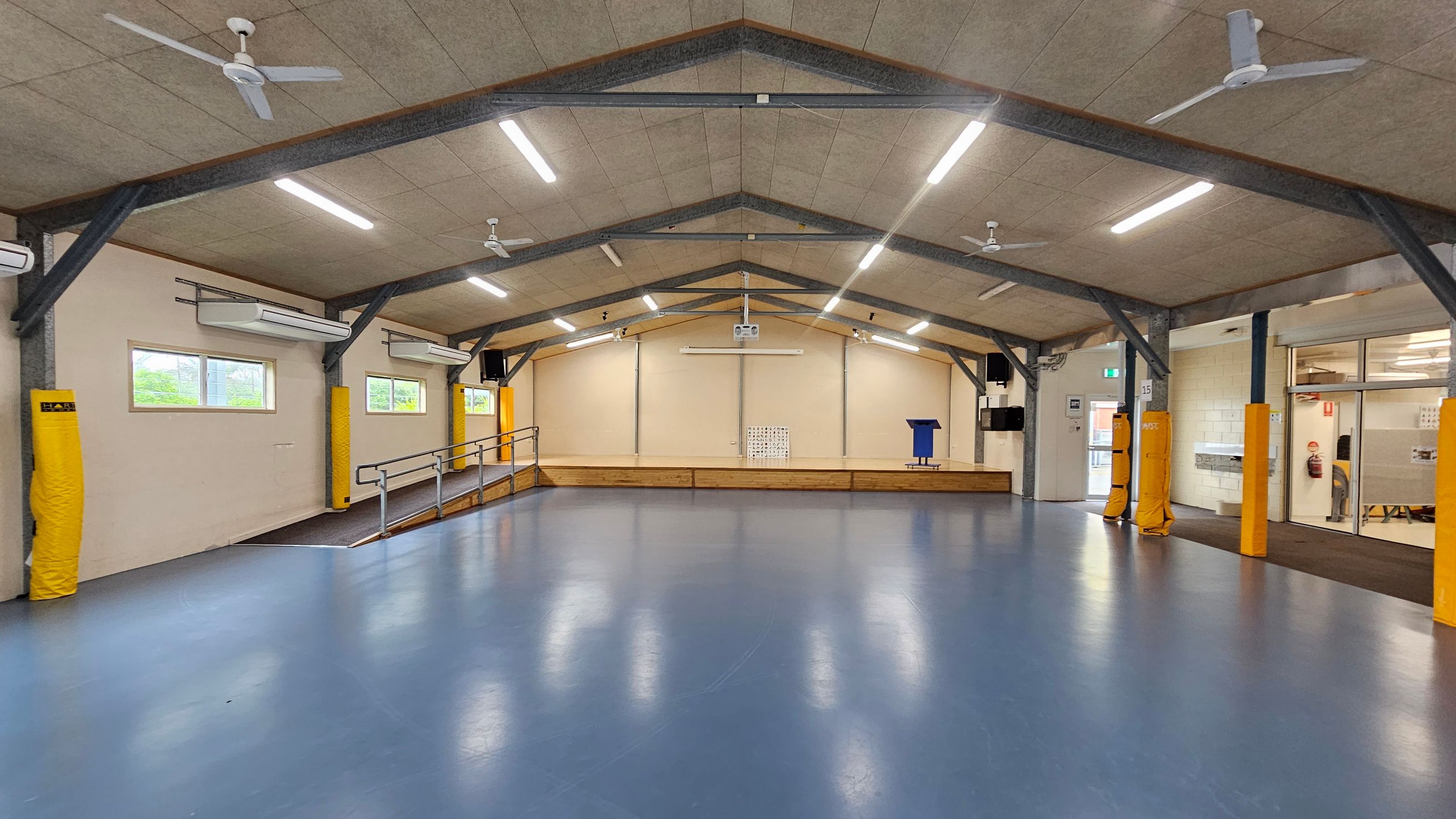A spacious indoor hall with a blue floor and a high, sloped ceiling featuring exposed beams and ceiling fans. The left side has windows with air conditioning units above, and yellow padding on the beams. The right side shows a glass entrance with a door and a fire extinguisher nearby. In the centre, a stage with a ramp, a blue lectern, and a projector screen is visible. The ceiling has multiple fluorescent lights. A spacious indoor hall with a blue floor and a high, sloped ceiling featuring exposed beams and ceiling fans. The left side has windows with air conditioning units above, and yellow padding on the beams. The right side shows a glass entrance with a door and a fire extinguisher nearby. In the centre, a stage with a ramp, a blue lectern, and a projector screen is visible. The ceiling has multiple fluorescent lights.