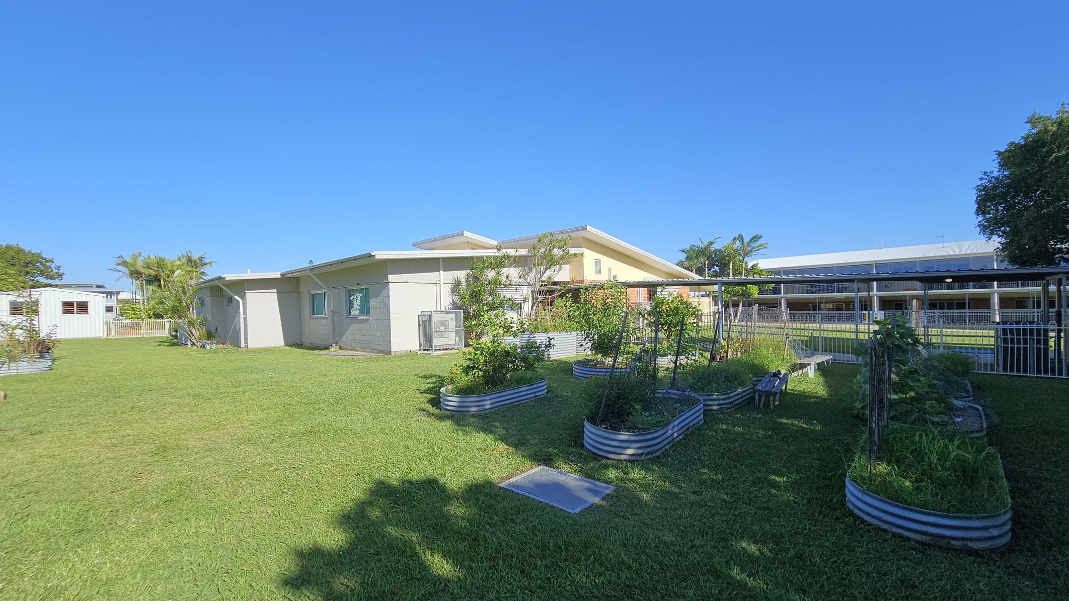 A grassy area with raised garden beds filled with plants is in the foreground. On the left, a light-coloured building with a sloped roof is visible, surrounded by a few small trees and shrubs. In the background, there is a larger building with a covered walkway and a metal fence. The sky is clear and blue. A grassy area with raised garden beds filled with plants is in the foreground. On the left, a light-coloured building with a sloped roof is visible, surrounded by a few small trees and shrubs. In the background, there is a larger building with a covered walkway and a metal fence. The sky is clear and blue.