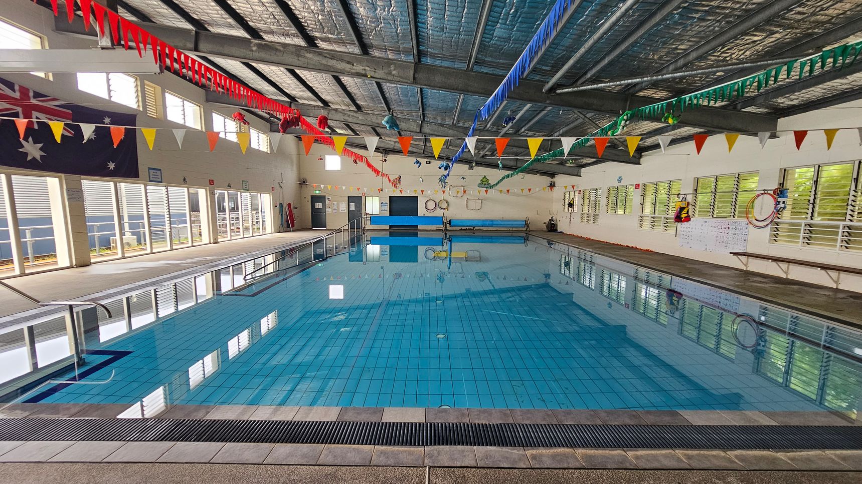 Indoor swimming pool with clear blue water, surrounded by a tiled deck. The ceiling is metal with exposed beams, adorned with colourful triangular bunting. On the left, large windows with louvres and an Australian flag. On the right, more windows and a wall with life rings and a notice board. The pool has a metal handrail on the left side. The floor is tiled, and the ceiling has insulation visible. Indoor swimming pool with clear blue water, surrounded by a tiled deck. The ceiling is metal with exposed beams, adorned with colourful triangular bunting. On the left, large windows with louvres and an Australian flag. On the right, more windows and a wall with life rings and a notice board. The pool has a metal handrail on the left side. The floor is tiled, and the ceiling has insulation visible.