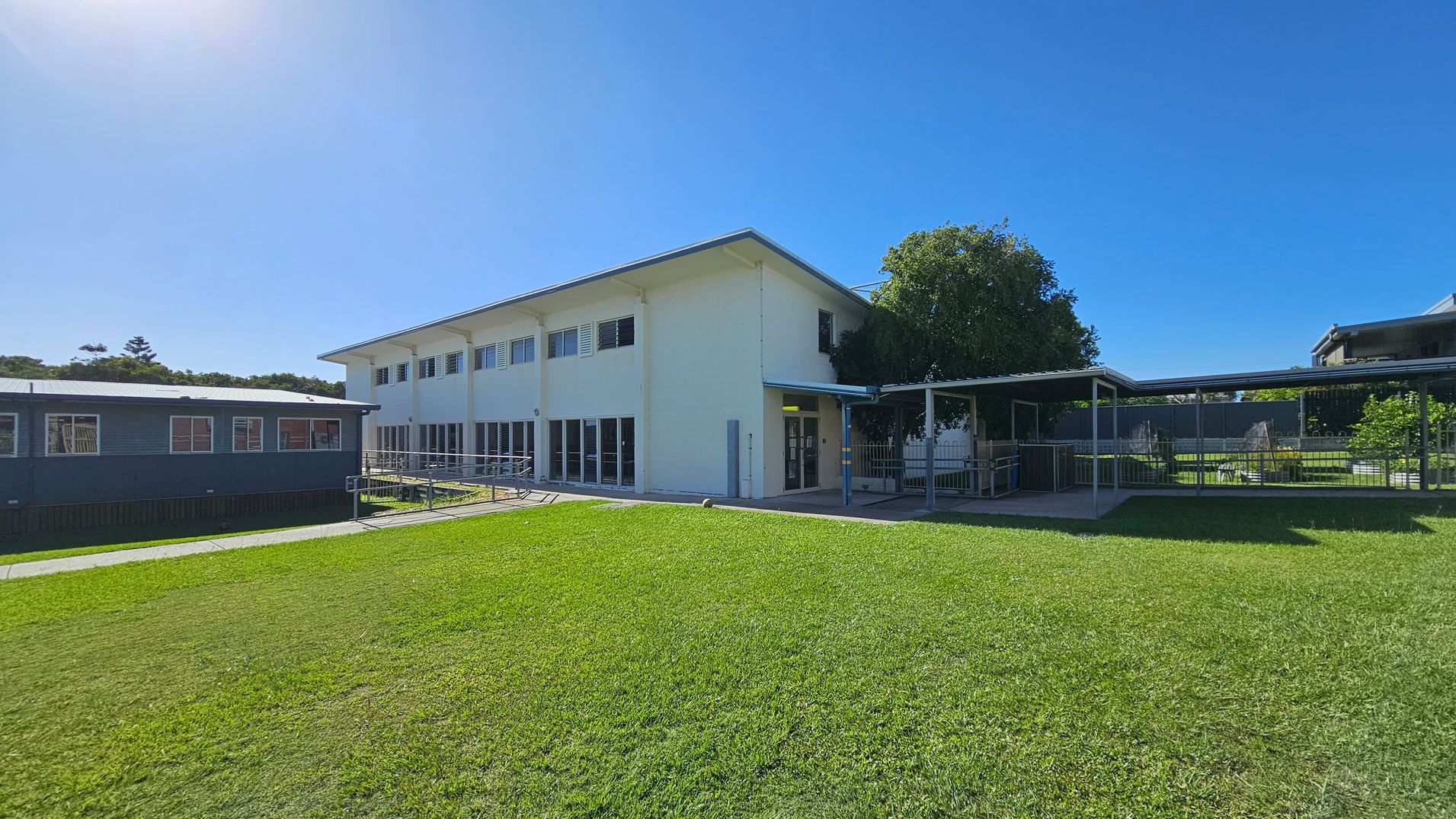 A two-storey white building with large windows on the right, connected to a blue single-storey structure on the left. A covered walkway extends from the main building, leading to a fenced area with greenery. The foreground features a well-maintained grassy lawn. A tree partially shades the entrance area. The sky is clear and blue. A two-storey white building with large windows on the right, connected to a blue single-storey structure on the left. A covered walkway extends from the main building, leading to a fenced area with greenery. The foreground features a well-maintained grassy lawn. A tree partially shades the entrance area. The sky is clear and blue.
