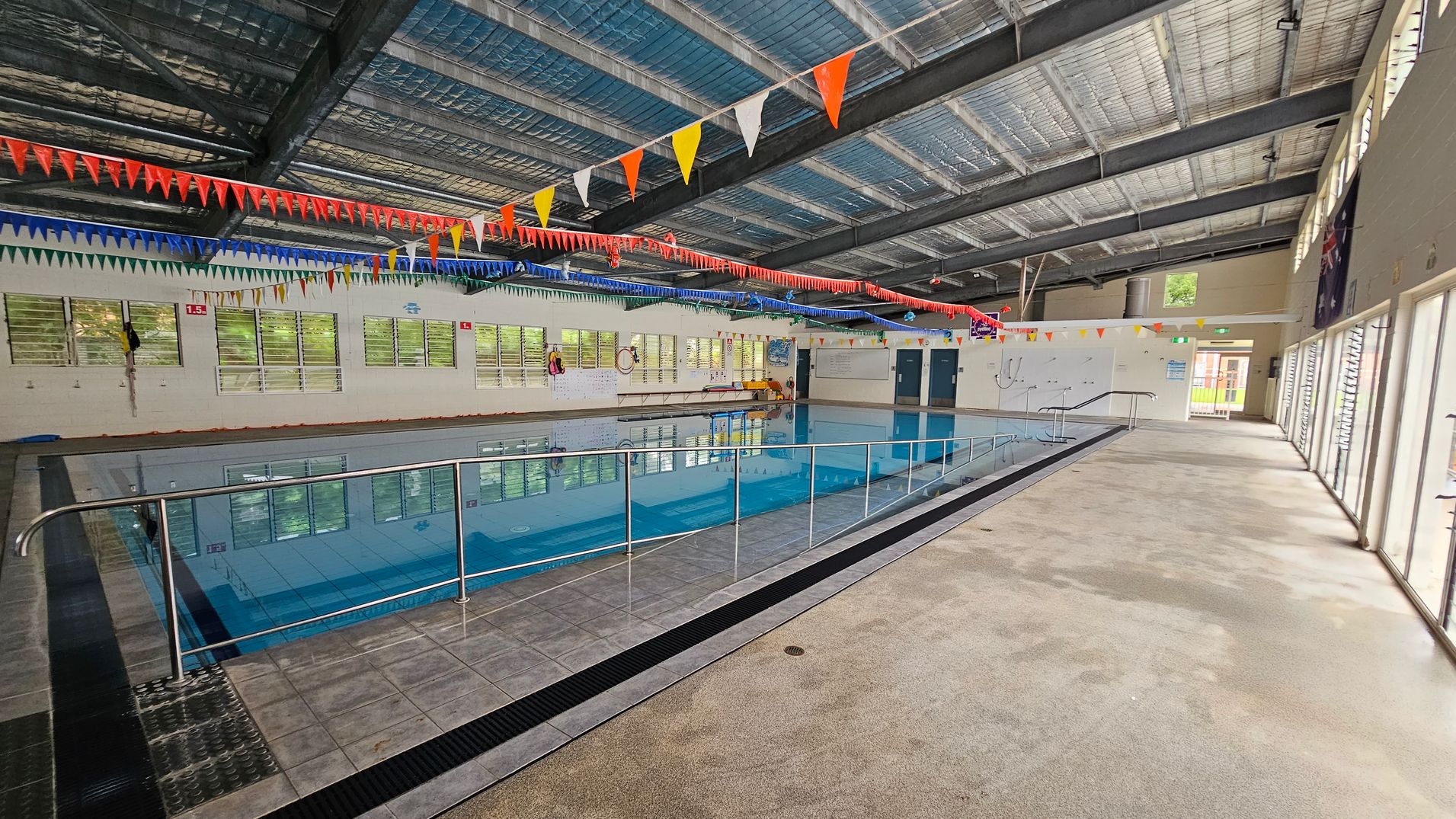 Indoor swimming pool with a tiled floor surrounding it. A metal railing runs along the pool's edge. The ceiling is made of exposed metal beams with colourful triangular flags hanging across. On the left, windows line the wall, allowing natural light in. The right side features large glass doors leading outside. The pool area is spacious, with a mix of tiled and carpeted flooring. Indoor swimming pool with a tiled floor surrounding it. A metal railing runs along the pool's edge. The ceiling is made of exposed metal beams with colourful triangular flags hanging across. On the left, windows line the wall, allowing natural light in. The right side features large glass doors leading outside. The pool area is spacious, with a mix of tiled and carpeted flooring.