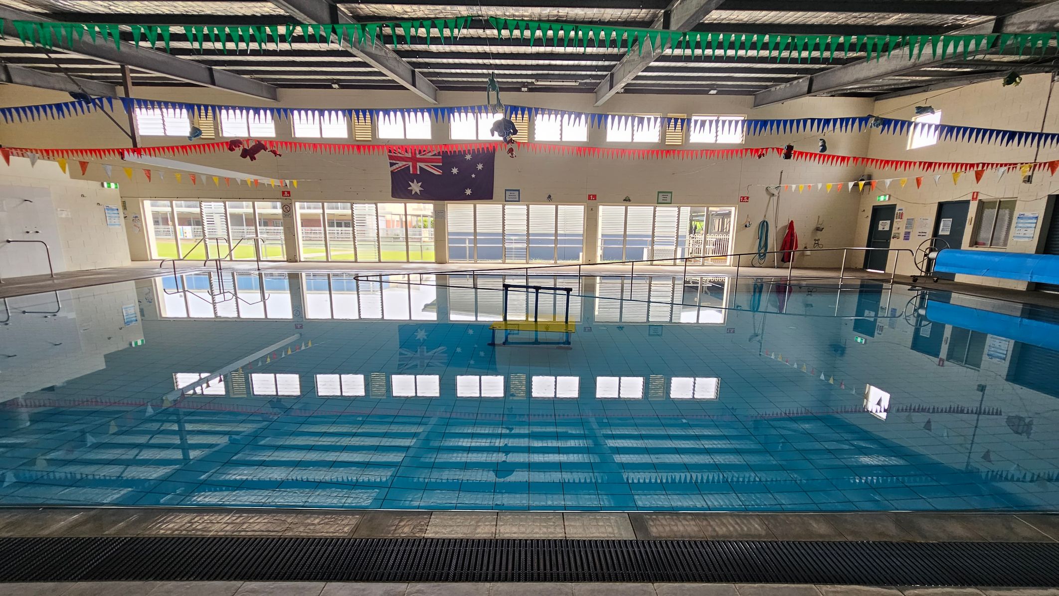 An indoor swimming pool with clear blue water. The ceiling has exposed beams and is adorned with colourful triangular bunting in green, blue, red, and yellow. Large windows line the far wall, allowing natural light to illuminate the space. An Australian flag is displayed on the wall. Pool equipment, such as a blue cover roller and a lifebuoy, is visible on the right. The floor around the pool is tiled, and a metal handrail is present at the pool's edge. An indoor swimming pool with clear blue water. The ceiling has exposed beams and is adorned with colourful triangular bunting in green, blue, red, and yellow. Large windows line the far wall, allowing natural light to illuminate the space. An Australian flag is displayed on the wall. Pool equipment, such as a blue cover roller and a lifebuoy, is visible on the right. The floor around the pool is tiled, and a metal handrail is present at the pool's edge.