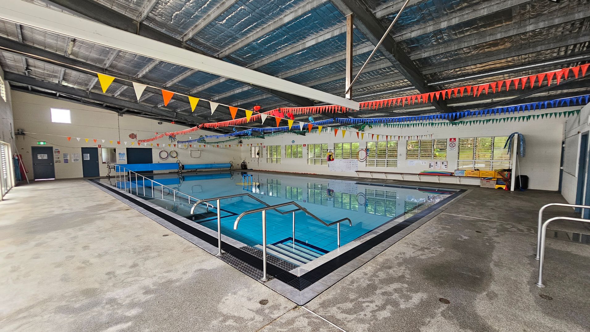 Indoor swimming pool with clear blue water, surrounded by a wet, textured floor. Metal handrails lead into the pool. Colourful triangular bunting hangs across the ceiling, which is metallic and industrial. On the left, a wall with doors and safety signs. On the right, windows with blinds, life rings, and pool equipment. The ceiling is high with exposed beams. Indoor swimming pool with clear blue water, surrounded by a wet, textured floor. Metal handrails lead into the pool. Colourful triangular bunting hangs across the ceiling, which is metallic and industrial. On the left, a wall with doors and safety signs. On the right, windows with blinds, life rings, and pool equipment. The ceiling is high with exposed beams.
