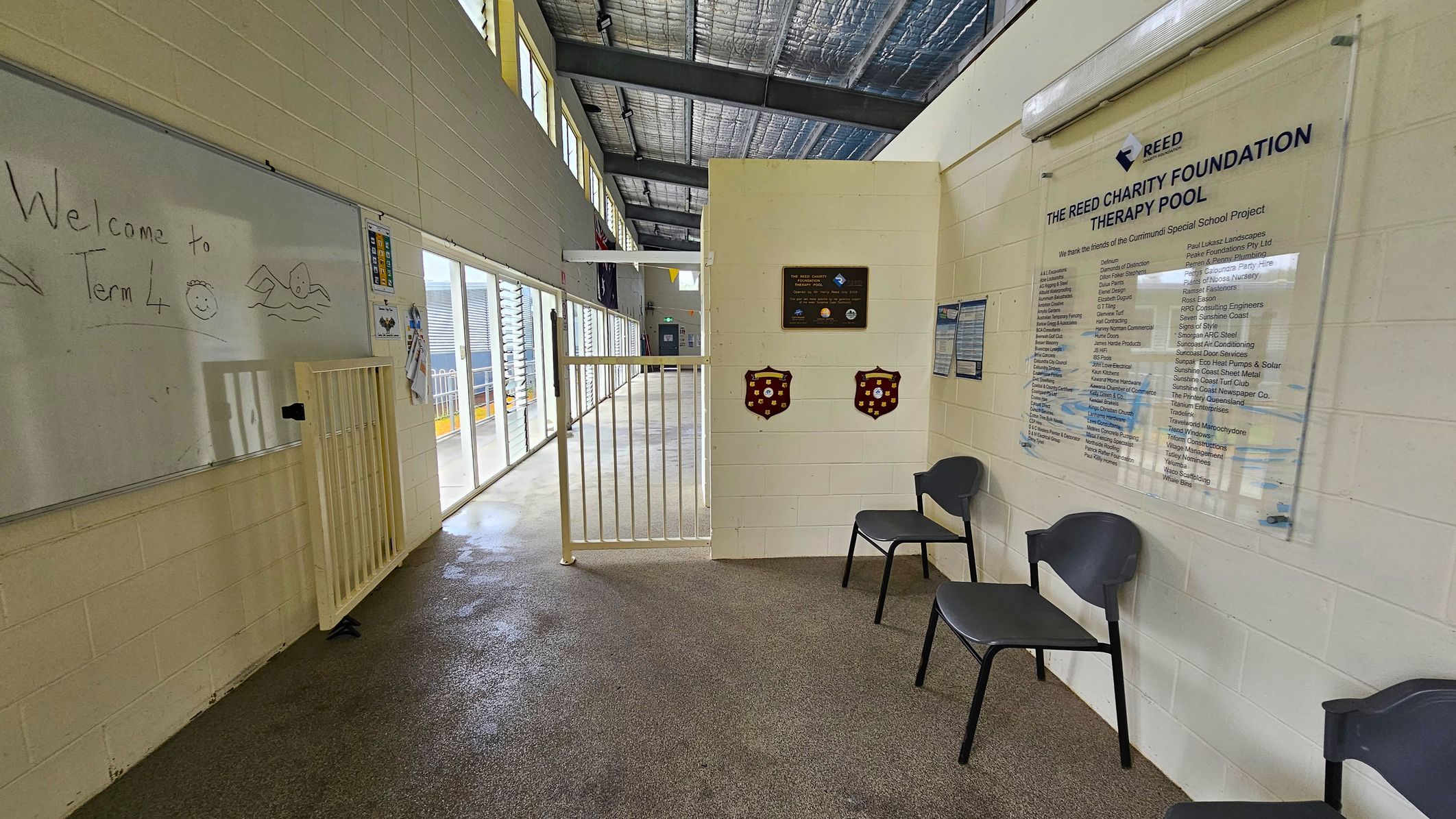 A hallway with a whiteboard on the left displaying "Welcome to Term 4" and a drawing of a swimmer. Below the board is a small gate. The right side features a wall with a plaque and a large sign for "The Reed Charity Foundation Therapy Pool," listing various names. Two black chairs are placed against the wall. The floor is a mix of wet and dry areas, and the ceiling has exposed metal beams and insulation. Large windows on the left allow natural light to enter. A hallway with a whiteboard on the left displaying "Welcome to Term 4" and a drawing of a swimmer. Below the board is a small gate. The right side features a wall with a plaque and a large sign for "The Reed Charity Foundation Therapy Pool," listing various names. Two black chairs are placed against the wall. The floor is a mix of wet and dry areas, and the ceiling has exposed metal beams and insulation. Large windows on the left allow natural light to enter.
