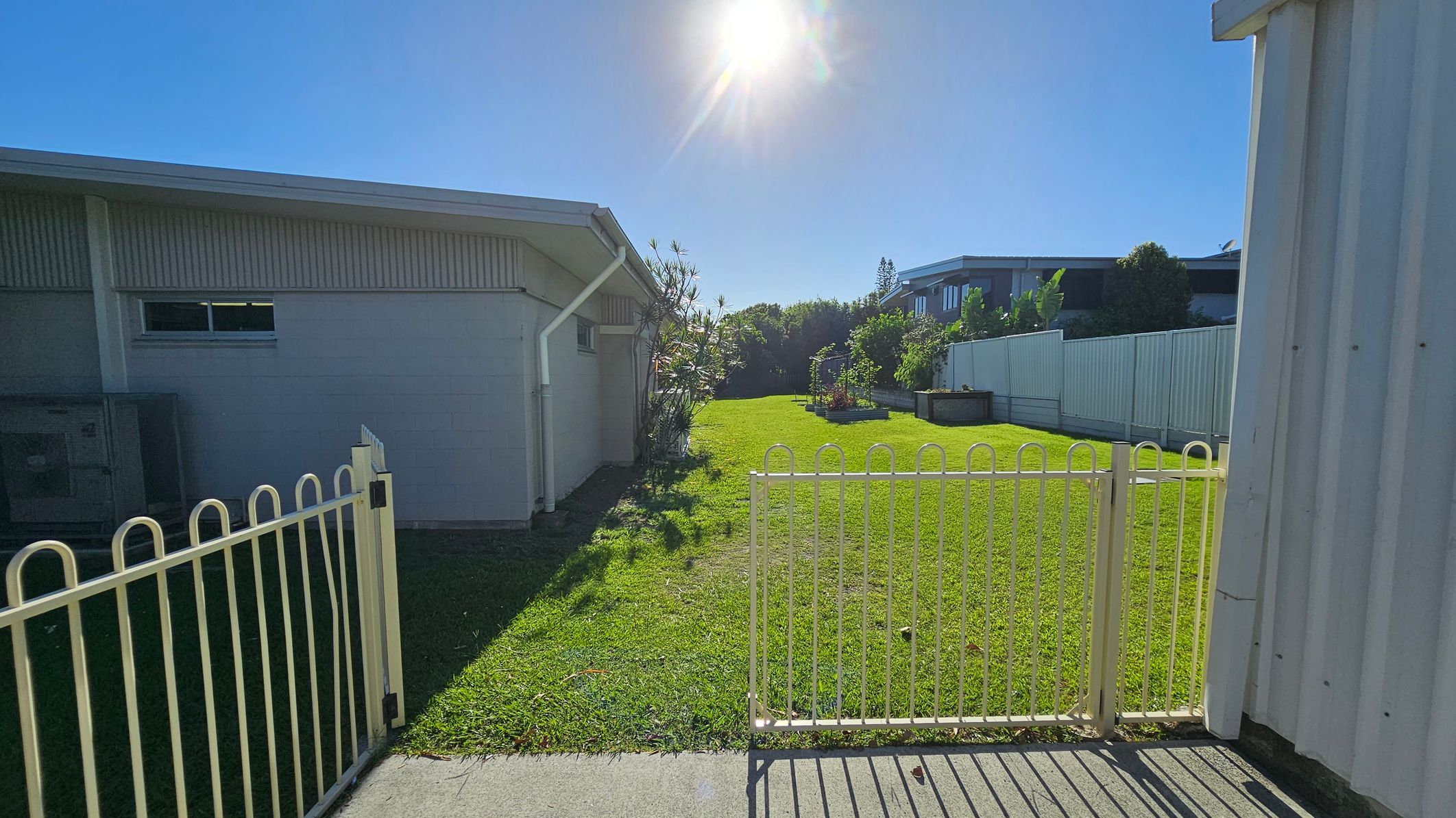 A grassy yard is bordered by a white fence, with a gate in the foreground. On the left, a building with a sloped roof and a small window is visible. On the right, another building with a corrugated exterior is partially seen. In the background, there are raised garden beds and a line of trees. The sun is shining brightly overhead, casting shadows on the grass. A grassy yard is bordered by a white fence, with a gate in the foreground. On the left, a building with a sloped roof and a small window is visible. On the right, another building with a corrugated exterior is partially seen. In the background, there are raised garden beds and a line of trees. The sun is shining brightly overhead, casting shadows on the grass.