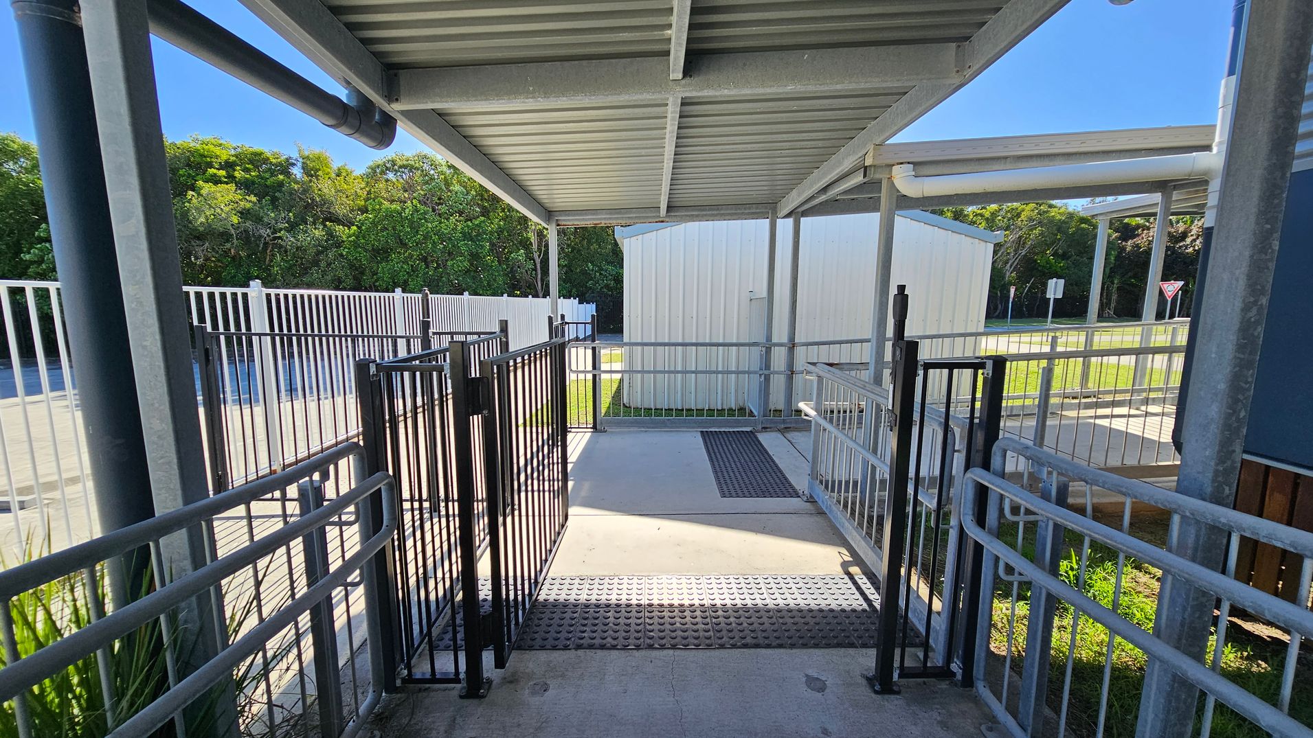 A covered walkway with a metal roof and railings on both sides. The floor is concrete with a textured mat in the centre. Two black gates are positioned along the path. In the background, there is a white building and a grassy area with trees. A metal fence runs parallel to the walkway on the left, and a yield sign is visible in the distance. A covered walkway with a metal roof and railings on both sides. The floor is concrete with a textured mat in the centre. Two black gates are positioned along the path. In the background, there is a white building and a grassy area with trees. A metal fence runs parallel to the walkway on the left, and a yield sign is visible in the distance.