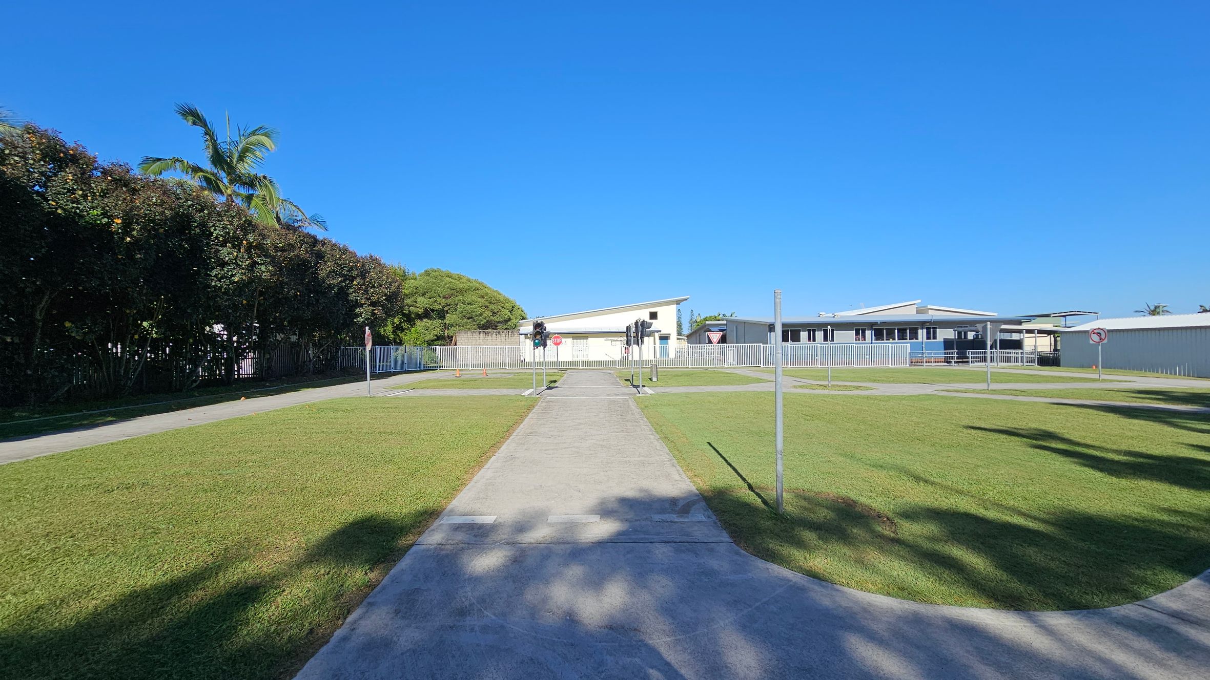 A paved path runs through the centre of a grassy area, leading to a building with a slanted roof in the background. On either side of the path are small traffic lights and signs, including a stop sign and a yield sign. To the left, a tall hedge with a palm tree is visible, while to the right, there are more buildings and a "no right turn" sign. Shadows from nearby trees are cast on the grass and path. A paved path runs through the centre of a grassy area, leading to a building with a slanted roof in the background. On either side of the path are small traffic lights and signs, including a stop sign and a yield sign. To the left, a tall hedge with a palm tree is visible, while to the right, there are more buildings and a "no right turn" sign. Shadows from nearby trees are cast on the grass and path.