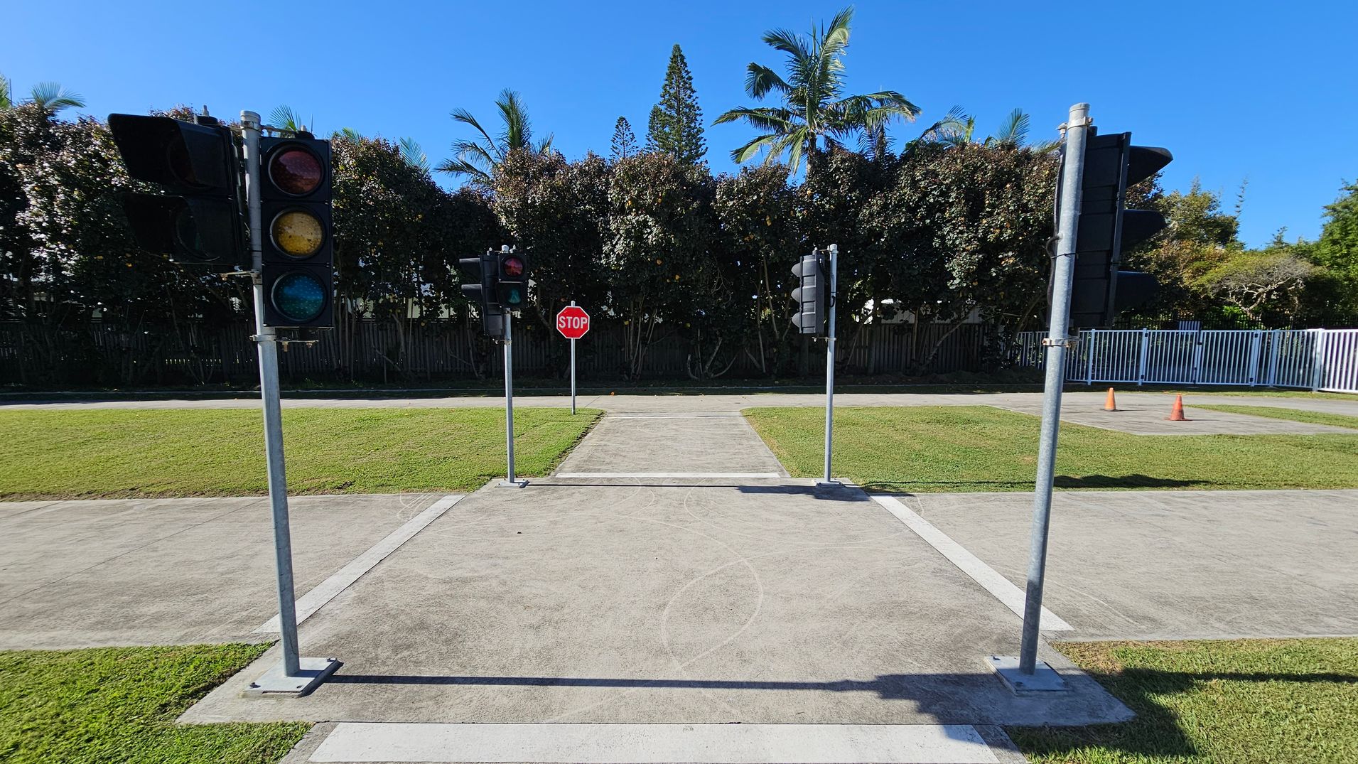 A small intersection with traffic lights on both sides and a stop sign in the centre. The ground is paved with concrete, surrounded by grass. In the background, there are tall trees and a white fence on the right. Two orange traffic cones are visible near the fence. The sky is clear and blue. A small intersection with traffic lights on both sides and a stop sign in the centre. The ground is paved with concrete, surrounded by grass. In the background, there are tall trees and a white fence on the right. Two orange traffic cones are visible near the fence. The sky is clear and blue.