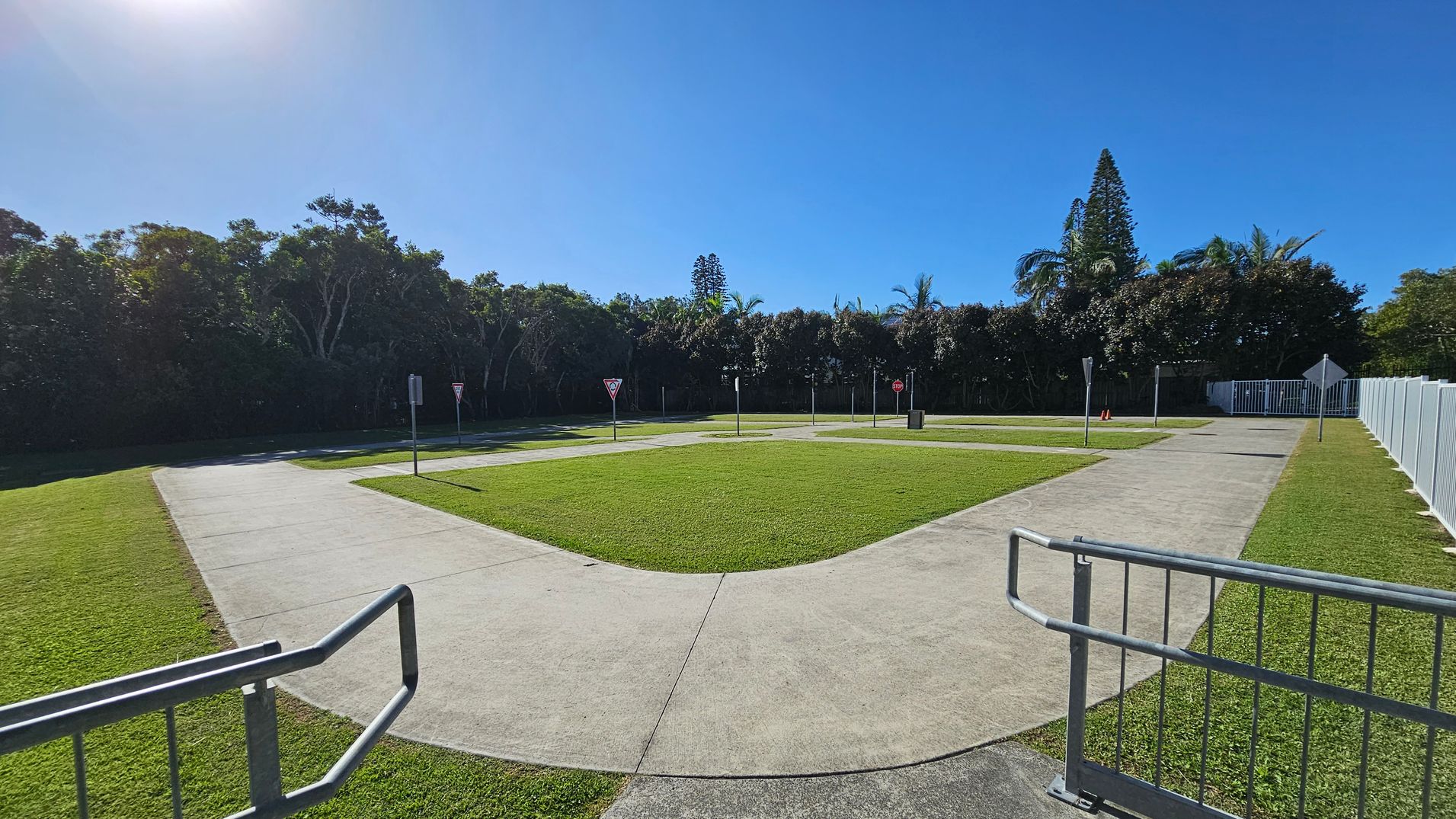 A small, fenced-in area with a paved path forming a loop around a grassy centre. Several traffic signs, including yield and stop signs, are placed along the path. The foreground features a metal railing. In the background, dense trees and tall coniferous trees are visible, with a clear blue sky above. The ground is covered with well-maintained grass. A small, fenced-in area with a paved path forming a loop around a grassy centre. Several traffic signs, including yield and stop signs, are placed along the path. The foreground features a metal railing. In the background, dense trees and tall coniferous trees are visible, with a clear blue sky above. The ground is covered with well-maintained grass.