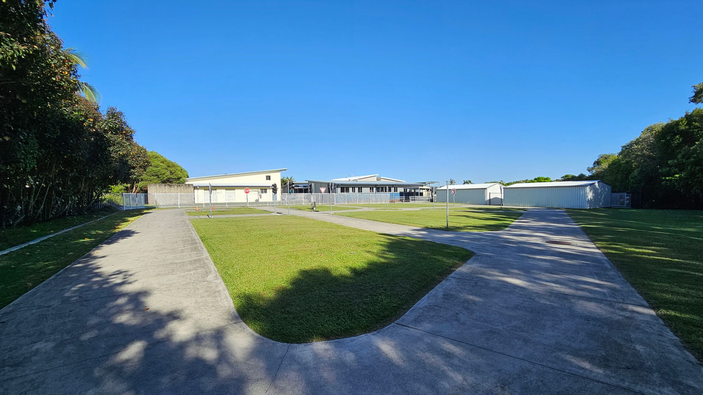 A paved path curves through a grassy area, leading towards a group of low, light-coloured buildings in the background. On the left, dense trees cast shadows on the path. On the right, more trees border the scene, with a white fence visible near the buildings. The sky is clear and blue. A paved path curves through a grassy area, leading towards a group of low, light-coloured buildings in the background. On the left, dense trees cast shadows on the path. On the right, more trees border the scene, with a white fence visible near the buildings. The sky is clear and blue.