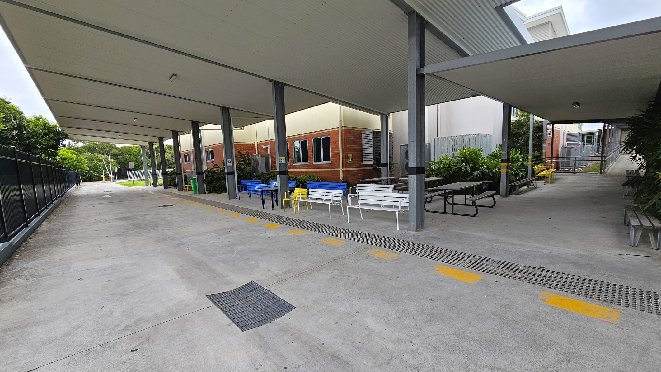 A covered walkway with a concrete floor and a metal ceiling supported by grey pillars. On the right, there are colourful benches in blue, yellow, and white, along with picnic tables. Behind the seating area, there are brick buildings with windows and some greenery. On the left, a black metal fence runs alongside the path, with trees visible in the background. Yellow markings are painted on the ground near the seating area. A covered walkway with a concrete floor and a metal ceiling supported by grey pillars. On the right, there are colourful benches in blue, yellow, and white, along with picnic tables. Behind the seating area, there are brick buildings with windows and some greenery. On the left, a black metal fence runs alongside the path, with trees visible in the background. Yellow markings are painted on the ground near the seating area.