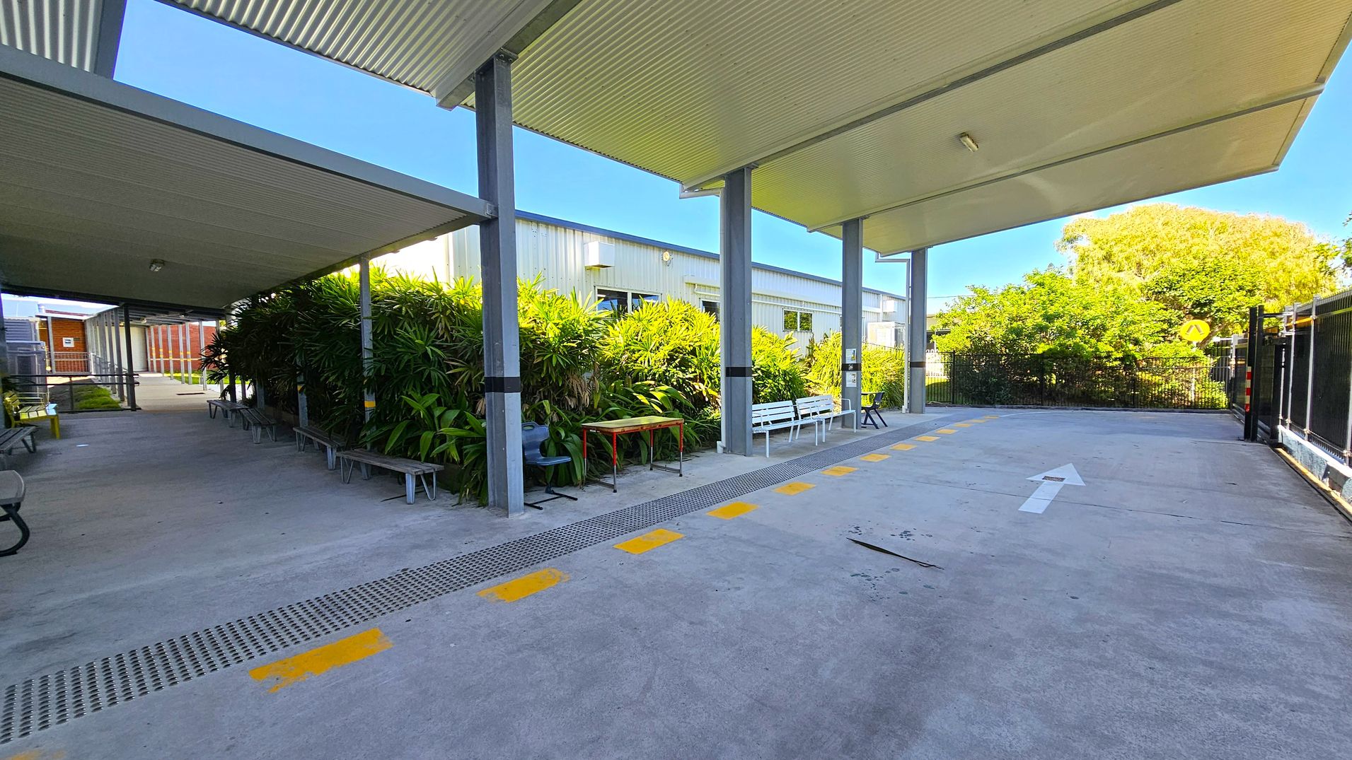 A covered outdoor walkway with a corrugated metal roof supported by metal beams. On the left, there are benches and a row of lush green plants. A white building with windows is visible behind the plants. On the right, a concrete path with a white arrow and yellow markings leads towards a fenced area with greenery. The ground is concrete, and the ceiling is metal. A covered outdoor walkway with a corrugated metal roof supported by metal beams. On the left, there are benches and a row of lush green plants. A white building with windows is visible behind the plants. On the right, a concrete path with a white arrow and yellow markings leads towards a fenced area with greenery. The ground is concrete, and the ceiling is metal.