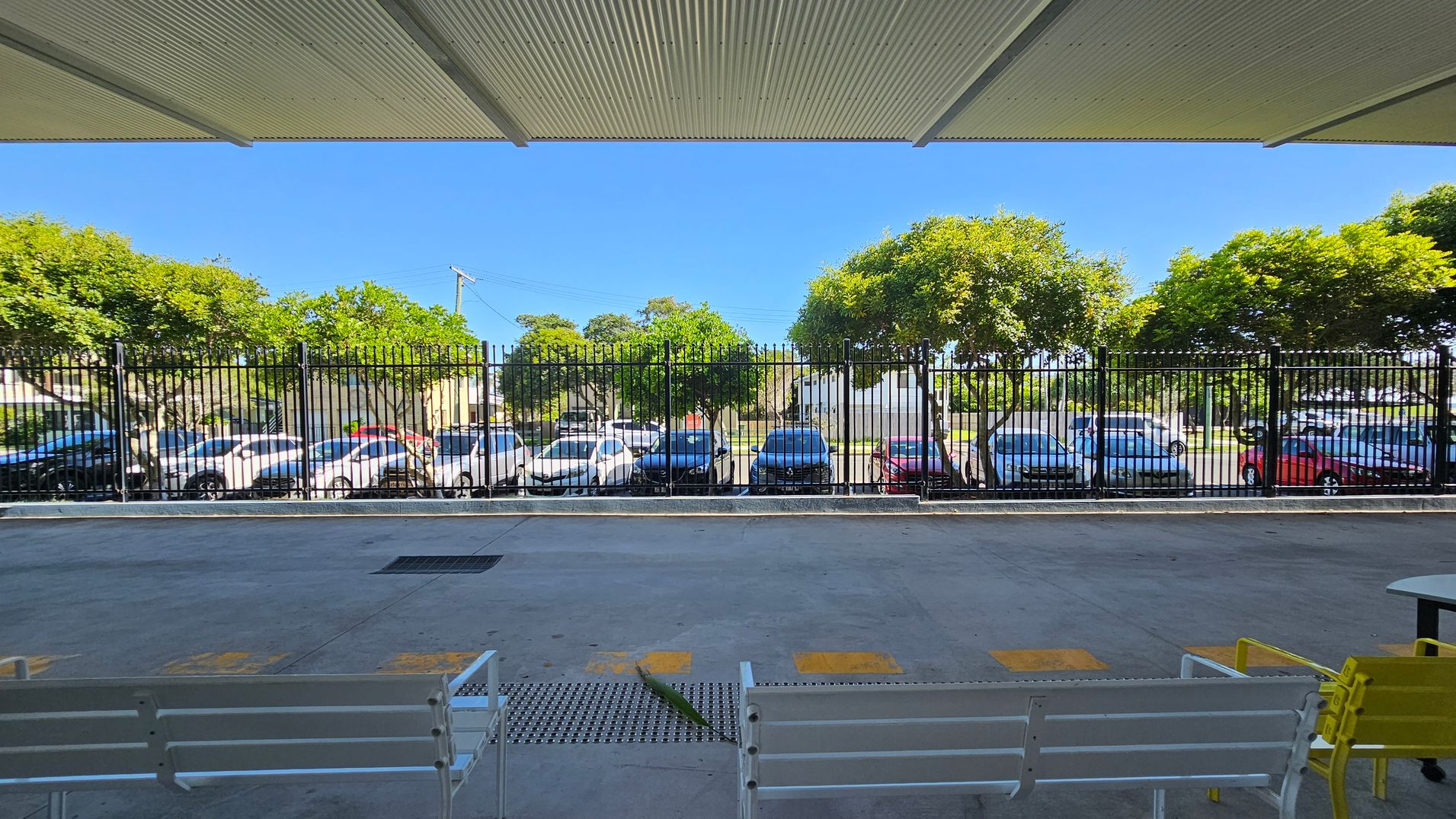 A covered area with a corrugated metal ceiling and a concrete floor. In the foreground, there are white benches and a yellow chair. Beyond a black metal fence, several parked cars are visible, including white, blue, and red vehicles. In the background, there are green trees and a clear blue sky. A covered area with a corrugated metal ceiling and a concrete floor. In the foreground, there are white benches and a yellow chair. Beyond a black metal fence, several parked cars are visible, including white, blue, and red vehicles. In the background, there are green trees and a clear blue sky.