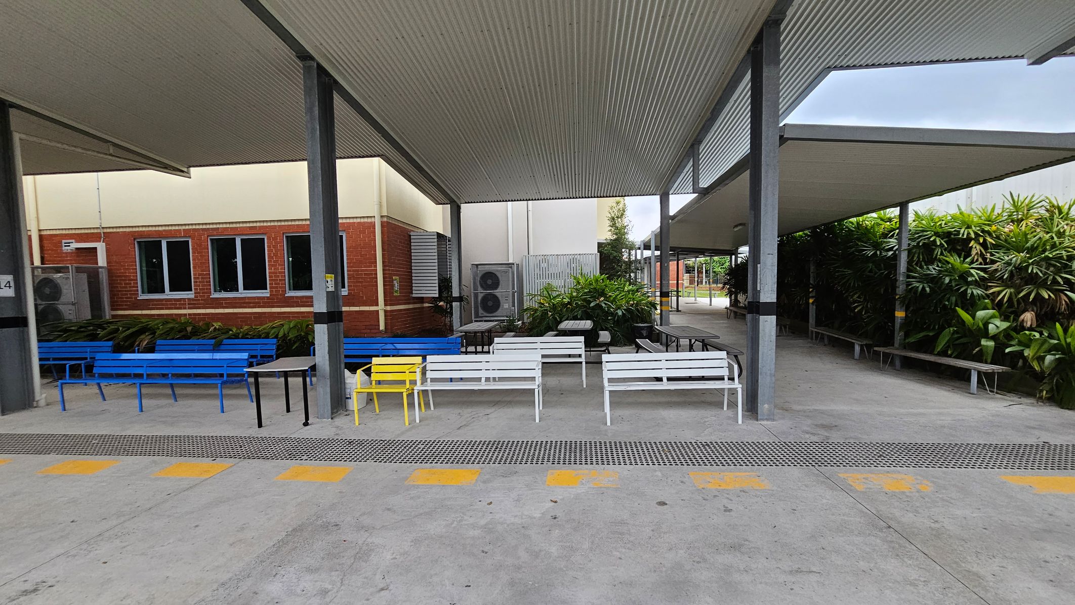 A covered outdoor area with a corrugated metal roof supported by metal beams. On the left, there are blue benches against a brick wall with windows and air conditioning units. In the centre, white benches and a yellow chair are arranged around tables. On the right, more benches are near lush green plants. The floor is concrete with yellow markings, and the ceiling is made of corrugated metal. A covered outdoor area with a corrugated metal roof supported by metal beams. On the left, there are blue benches against a brick wall with windows and air conditioning units. In the centre, white benches and a yellow chair are arranged around tables. On the right, more benches are near lush green plants. The floor is concrete with yellow markings, and the ceiling is made of corrugated metal.