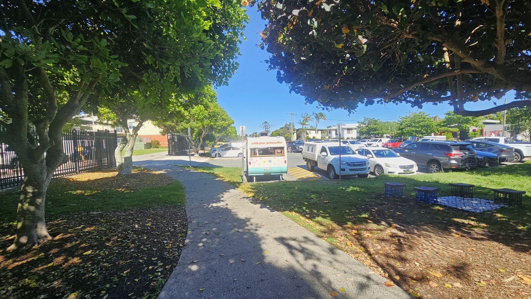 A pathway curves through a park area, bordered by trees with dense green foliage. On the left, a black metal fence runs alongside the path. In the centre, a small ice cream van is parked near the road. To the right, several parked cars are visible in a parking area. The ground is a mix of grass and mulch, with some fallen leaves. Milk crates are arranged on the grass, possibly as makeshift seating. A pathway curves through a park area, bordered by trees with dense green foliage. On the left, a black metal fence runs alongside the path. In the centre, a small ice cream van is parked near the road. To the right, several parked cars are visible in a parking area. The ground is a mix of grass and mulch, with some fallen leaves. Milk crates are arranged on the grass, possibly as makeshift seating.