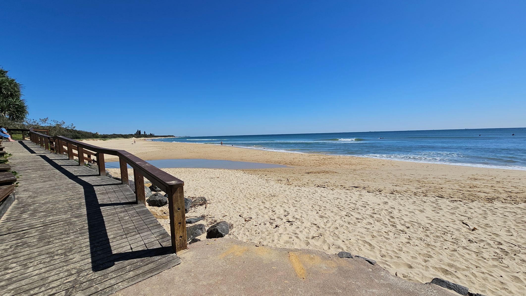 A wooden boardwalk with a railing runs along the left side, leading towards the background. On the right, a sandy beach stretches out to meet the ocean, with gentle waves lapping at the shore. In the distance, a few people are visible near the water. The foreground shows a sandy path with some rocks. Vegetation is visible on the far left, and the sky is clear and blue. A wooden boardwalk with a railing runs along the left side, leading towards the background. On the right, a sandy beach stretches out to meet the ocean, with gentle waves lapping at the shore. In the distance, a few people are visible near the water. The foreground shows a sandy path with some rocks. Vegetation is visible on the far left, and the sky is clear and blue.