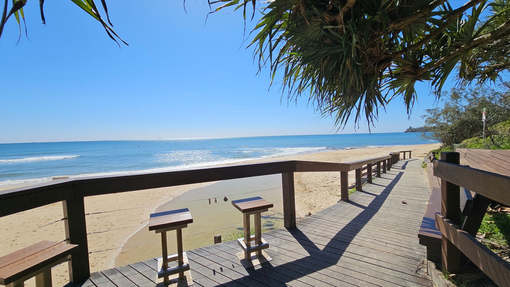 A wooden boardwalk runs along a sandy beach, with two stools positioned at a railing overlooking the ocean. The sea is calm with gentle waves. On the right, the boardwalk extends into the distance, bordered by greenery. Overhead, leafy branches hang down, partially shading the area. The beach stretches out to the left, meeting the blue water under a clear sky. A wooden boardwalk runs along a sandy beach, with two stools positioned at a railing overlooking the ocean. The sea is calm with gentle waves. On the right, the boardwalk extends into the distance, bordered by greenery. Overhead, leafy branches hang down, partially shading the area. The beach stretches out to the left, meeting the blue water under a clear sky.