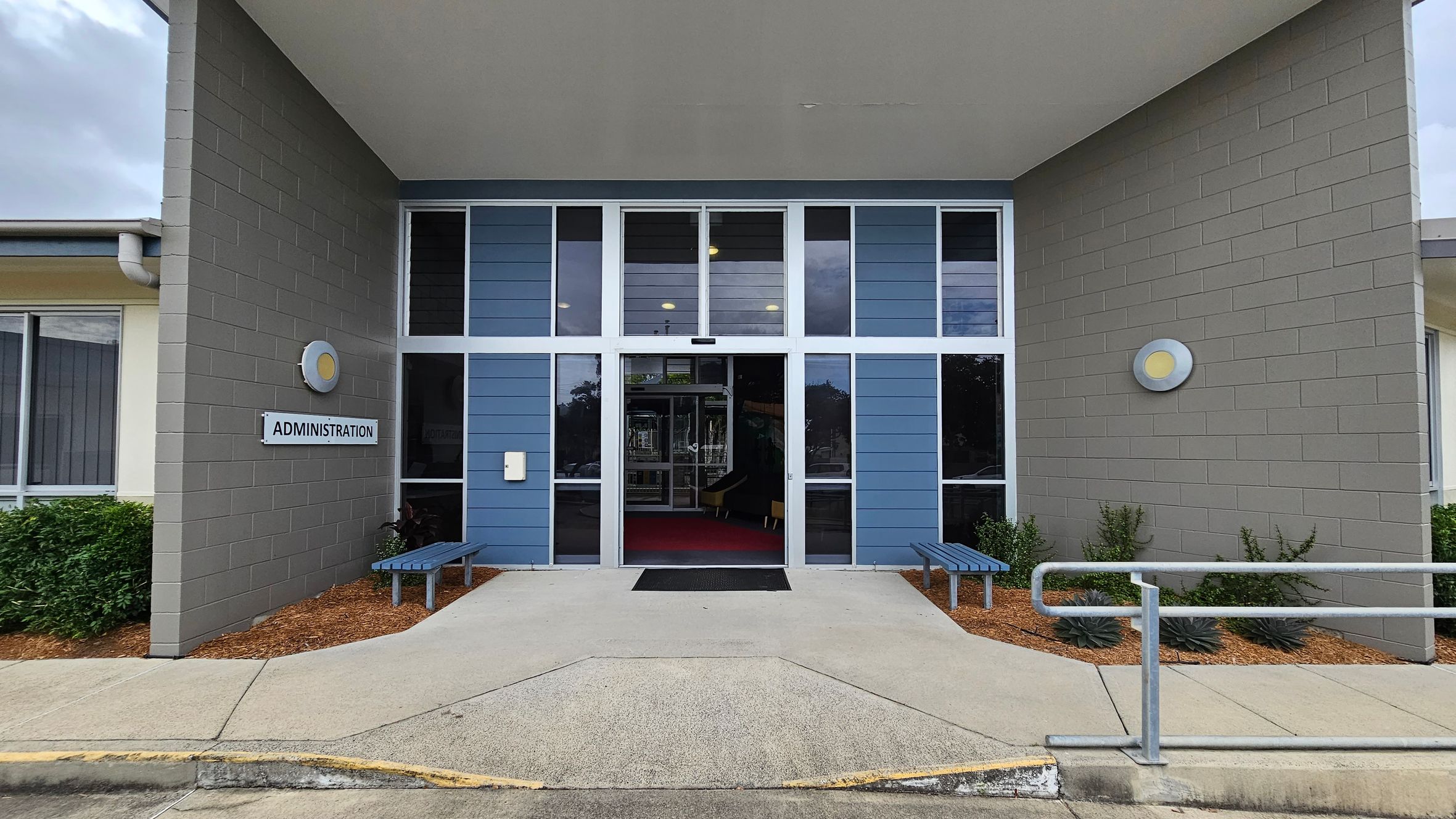 Entrance to a building with a sign reading "Administration" on the left wall. The facade features grey brick walls and blue panelling with large glass windows. Two circular lights are mounted on each side. A pathway leads to the entrance, flanked by two blue benches. A metal railing is on the right, and there are small shrubs planted along the walls. The ceiling is flat and extends over the entrance area. Entrance to a building with a sign reading "Administration" on the left wall. The facade features grey brick walls and blue panelling with large glass windows. Two circular lights are mounted on each side. A pathway leads to the entrance, flanked by two blue benches. A metal railing is on the right, and there are small shrubs planted along the walls. The ceiling is flat and extends over the entrance area.