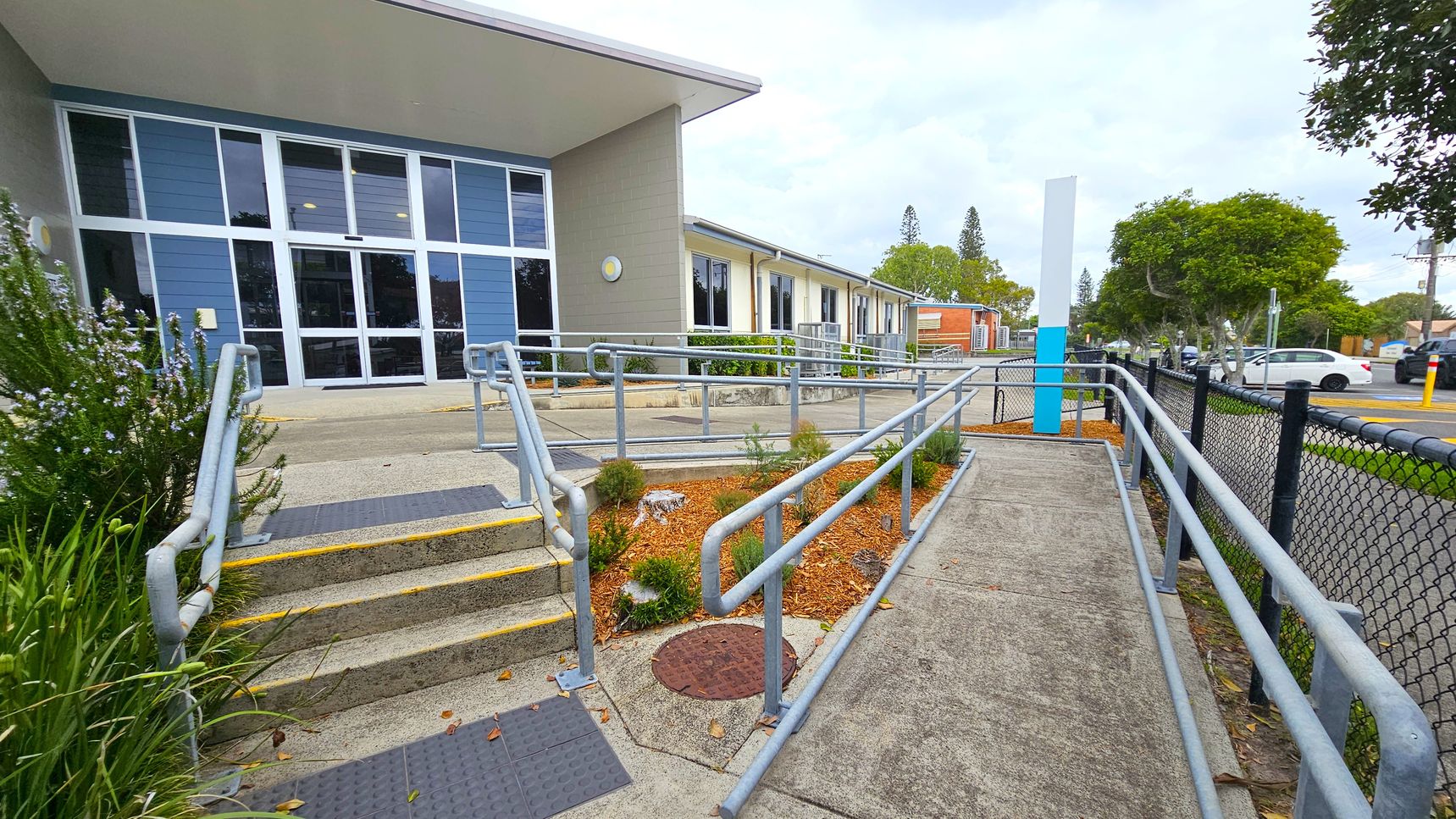 A building with large blue and grey windows on the left, featuring a ramp and steps with metal railings leading up to the entrance. The foreground has a landscaped area with small plants and mulch. On the right, a black chain-link fence runs alongside a pavement, with trees and parked cars visible in the background. The ground is concrete, and the ceiling of the entrance area is flat and white. A building with large blue and grey windows on the left, featuring a ramp and steps with metal railings leading up to the entrance. The foreground has a landscaped area with small plants and mulch. On the right, a black chain-link fence runs alongside a pavement, with trees and parked cars visible in the background. The ground is concrete, and the ceiling of the entrance area is flat and white.