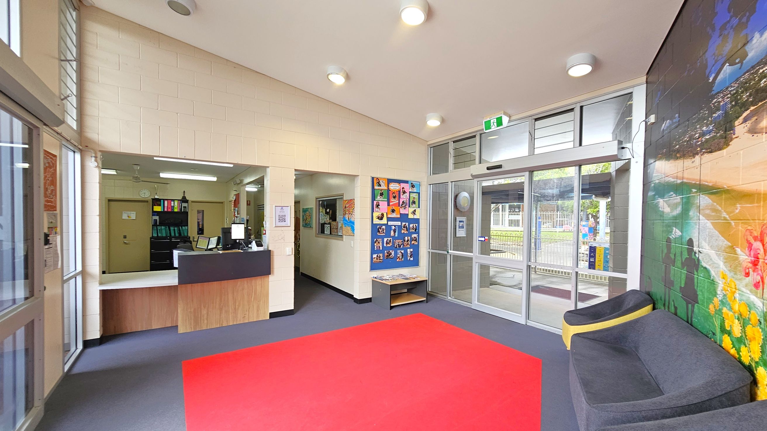 A reception area with a counter on the left, featuring a computer and office supplies. Behind the counter, there are shelves with colourful binders. In the centre, a wall displays a bulletin board with photos and artwork. To the right, a glass door leads outside, with a mural of a beach scene on the adjacent wall. A grey and yellow seating area is positioned near the mural. The floor is covered with a red carpet, and the ceiling has several round lights. A reception area with a counter on the left, featuring a computer and office supplies. Behind the counter, there are shelves with colourful binders. In the centre, a wall displays a bulletin board with photos and artwork. To the right, a glass door leads outside, with a mural of a beach scene on the adjacent wall. A grey and yellow seating area is positioned near the mural. The floor is covered with a red carpet, and the ceiling has several round lights.