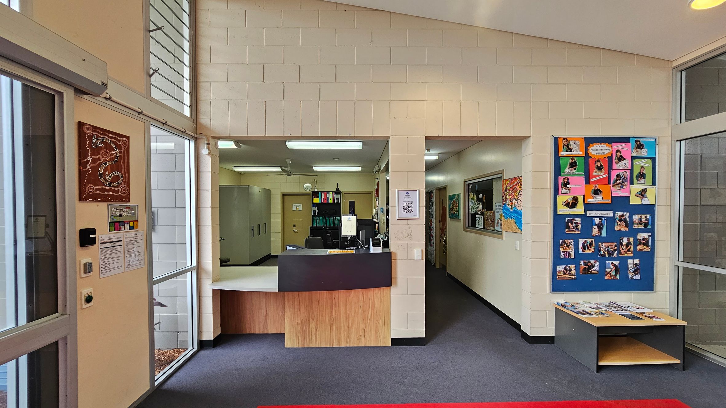 A reception area with a counter in the centre, featuring a computer and office supplies. To the left, there are large windows with a wall-mounted artwork and notices. On the right, a colourful bulletin board displays various photos and papers. Below the board is a small table with brochures. The floor is carpeted in dark grey, and the ceiling has fluorescent lights. A reception area with a counter in the centre, featuring a computer and office supplies. To the left, there are large windows with a wall-mounted artwork and notices. On the right, a colourful bulletin board displays various photos and papers. Below the board is a small table with brochures. The floor is carpeted in dark grey, and the ceiling has fluorescent lights.