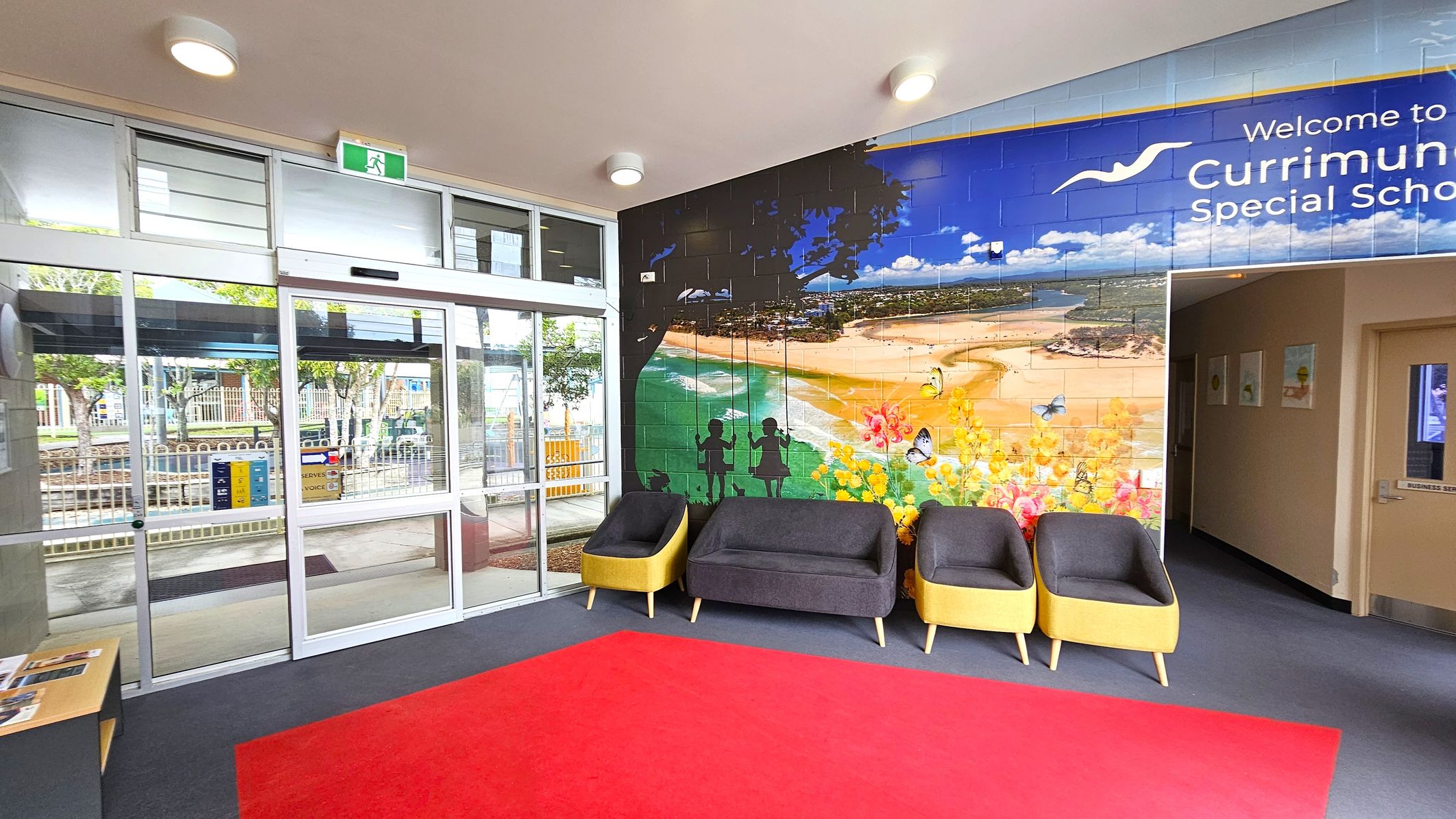 Entrance area with a glass sliding door on the left, leading outside. Above the door is an emergency exit sign. The right wall features a large mural depicting a beach scene with silhouettes of children on swings and colourful flowers. Below the mural are three chairs with grey cushions and yellow sides. The floor is covered with a red carpet, and the ceiling has round lights. A small table with brochures is near the door. Entrance area with a glass sliding door on the left, leading outside. Above the door is an emergency exit sign. The right wall features a large mural depicting a beach scene with silhouettes of children on swings and colourful flowers. Below the mural are three chairs with grey cushions and yellow sides. The floor is covered with a red carpet, and the ceiling has round lights. A small table with brochures is near the door.