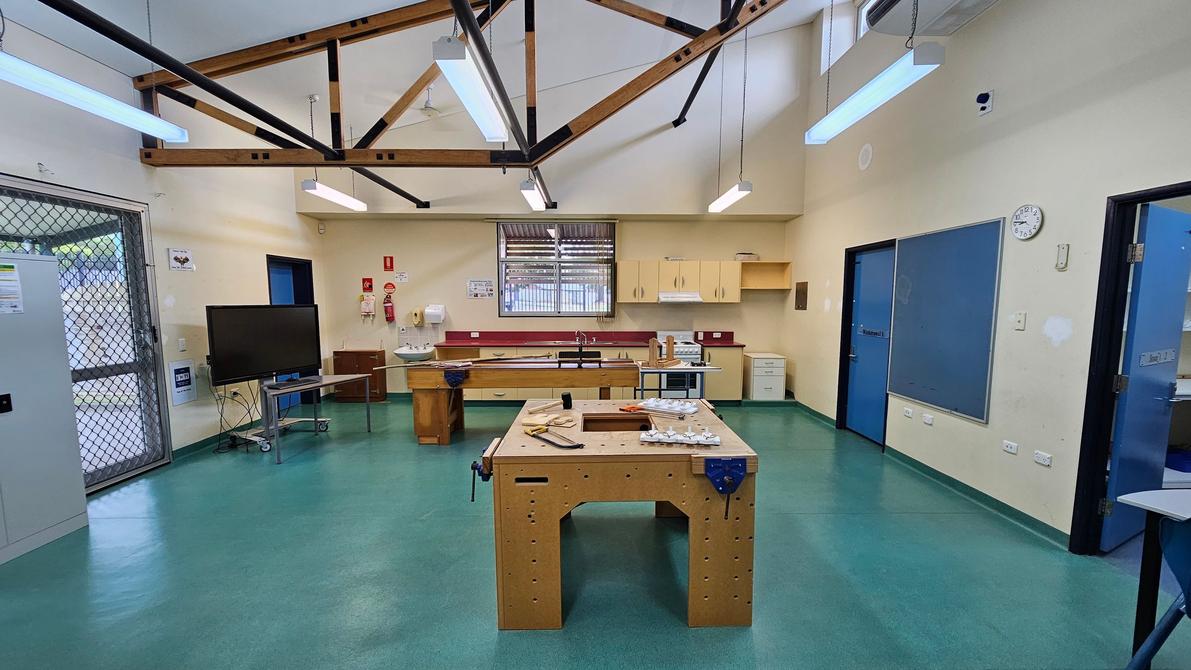 A workshop room with a green floor and high ceiling featuring exposed wooden beams. In the centre, a wooden workbench is equipped with tools and materials. The back wall has a window with a metal grille, surrounded by cabinets and a sink. On the left, a large TV on a stand is near a metal mesh door. The right side has blue doors and a clock on the wall. Fluorescent lights hang from the ceiling. A workshop room with a green floor and high ceiling featuring exposed wooden beams. In the centre, a wooden workbench is equipped with tools and materials. The back wall has a window with a metal grille, surrounded by cabinets and a sink. On the left, a large TV on a stand is near a metal mesh door. The right side has blue doors and a clock on the wall. Fluorescent lights hang from the ceiling.