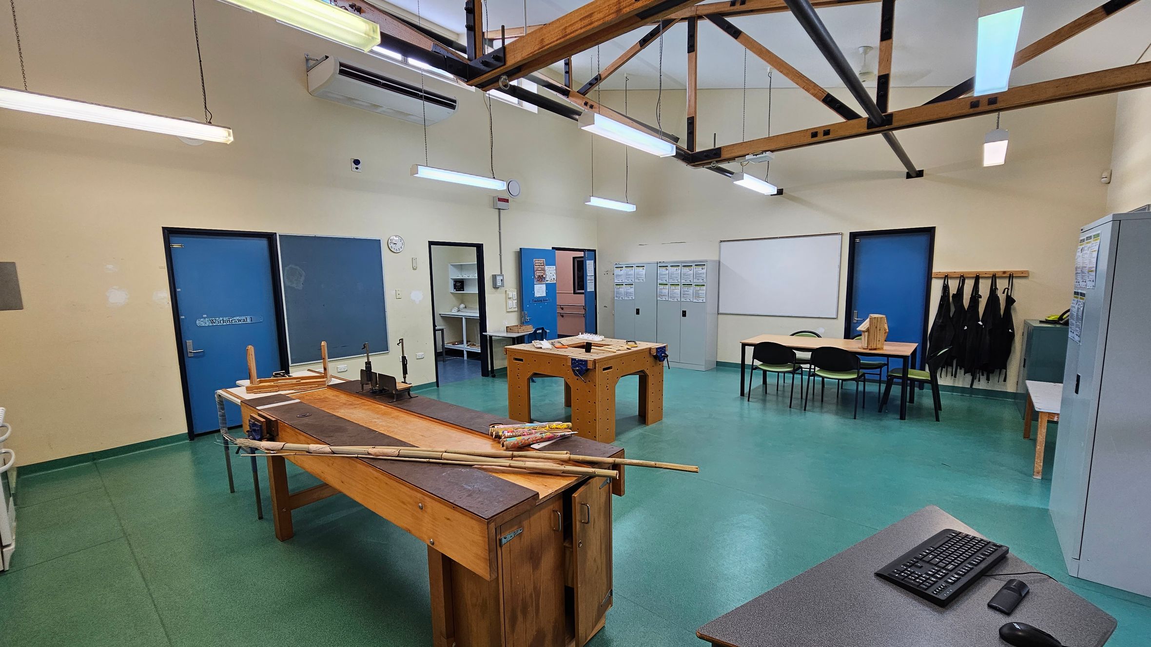 A workshop room with green flooring and a high ceiling featuring exposed wooden beams. In the foreground, a wooden workbench with tools and materials is visible. To the right, a grey table with a keyboard and mouse. The background has blue doors, a clock, and a whiteboard. Lockers and a table with chairs are on the right, with black aprons hanging on hooks. Fluorescent lights hang from the ceiling. A workshop room with green flooring and a high ceiling featuring exposed wooden beams. In the foreground, a wooden workbench with tools and materials is visible. To the right, a grey table with a keyboard and mouse. The background has blue doors, a clock, and a whiteboard. Lockers and a table with chairs are on the right, with black aprons hanging on hooks. Fluorescent lights hang from the ceiling.
