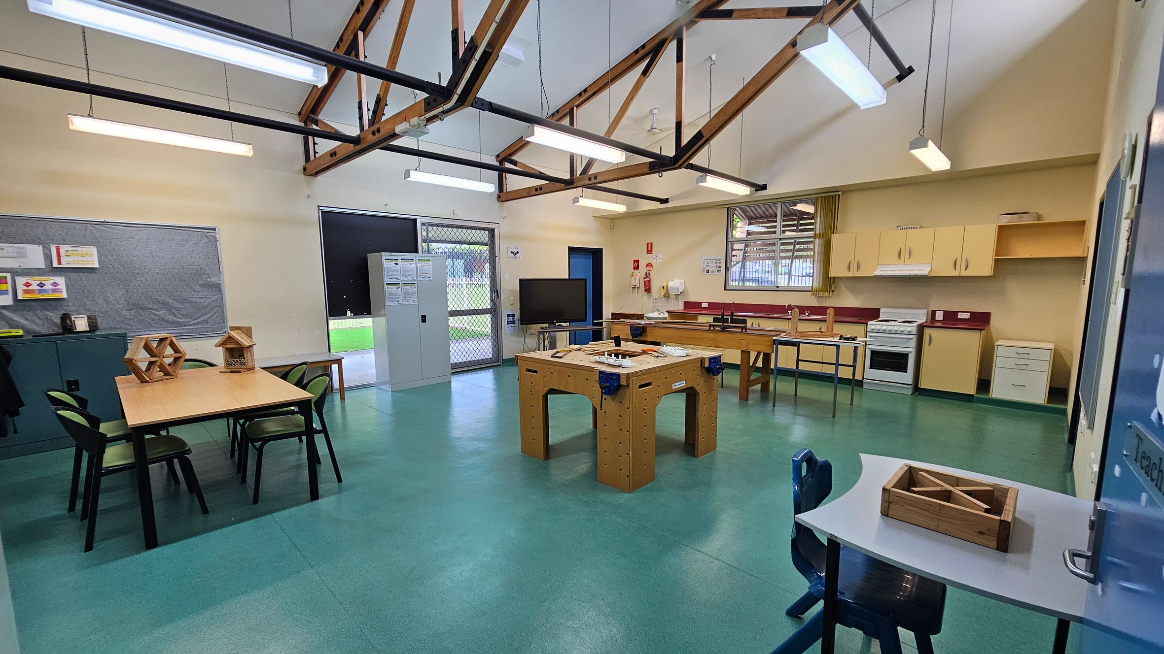 A classroom with a green floor and high ceiling featuring exposed wooden beams. On the left, there's a table with chairs and geometric wooden models. A bulletin board and storage cabinet are nearby. In the centre, a large wooden workbench is visible. The right side has a kitchen area with cabinets, a stove, and a sink. A small desk with a chair and wooden box is in the foreground. Fluorescent lights hang from the ceiling, and a door and windows are in the background. A classroom with a green floor and high ceiling featuring exposed wooden beams. On the left, there's a table with chairs and geometric wooden models. A bulletin board and storage cabinet are nearby. In the centre, a large wooden workbench is visible. The right side has a kitchen area with cabinets, a stove, and a sink. A small desk with a chair and wooden box is in the foreground. Fluorescent lights hang from the ceiling, and a door and windows are in the background.