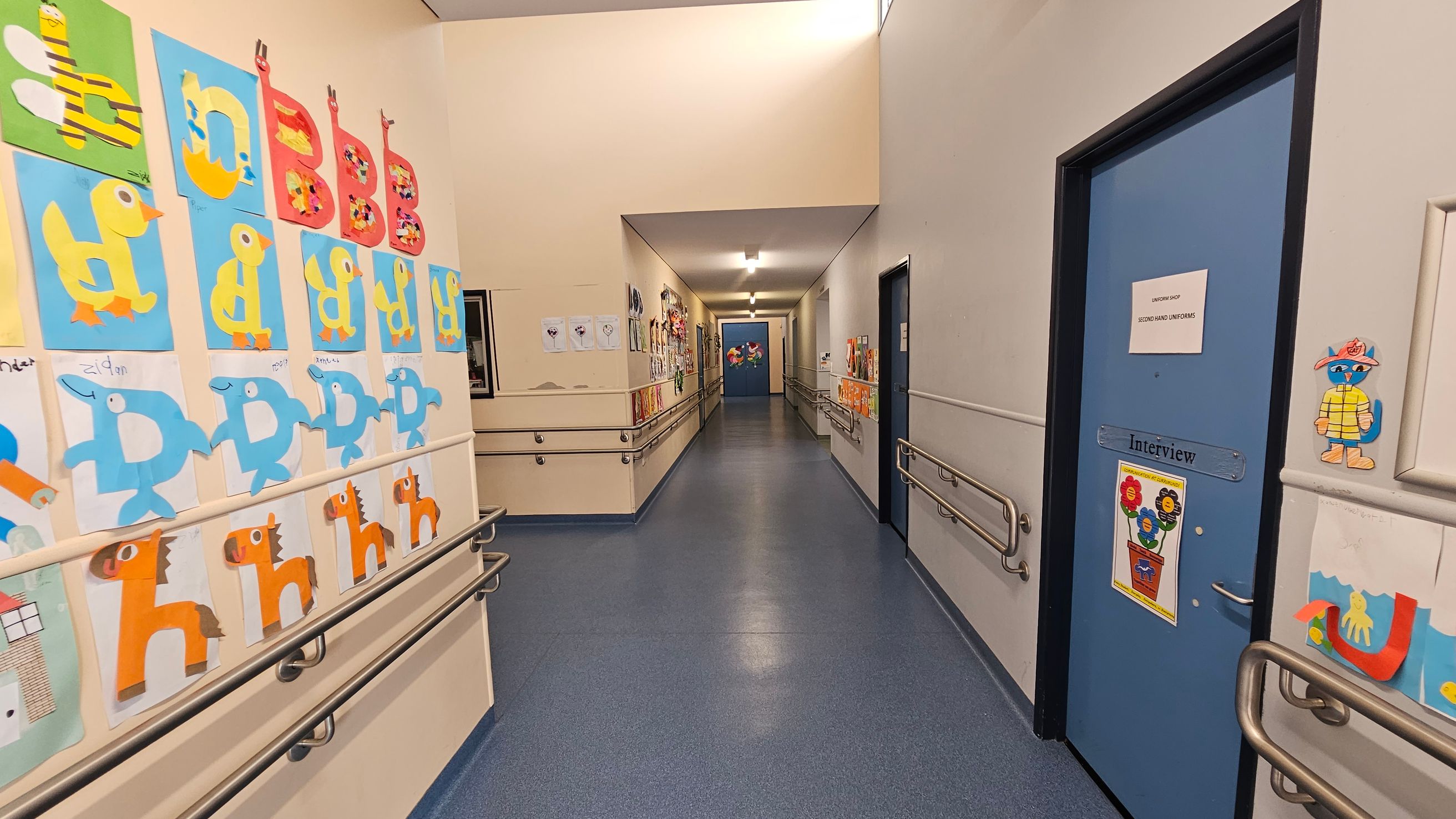 A corridor with blue flooring and beige walls. On the left, colourful children's artwork featuring animals and letters is displayed. On the right, a blue door with a sign reading "Interview" and more artwork is visible. Handrails run along both sides of the corridor. The ceiling is white with fluorescent lighting, and more artwork is visible further down the hallway. A corridor with blue flooring and beige walls. On the left, colourful children's artwork featuring animals and letters is displayed. On the right, a blue door with a sign reading "Interview" and more artwork is visible. Handrails run along both sides of the corridor. The ceiling is white with fluorescent lighting, and more artwork is visible further down the hallway.